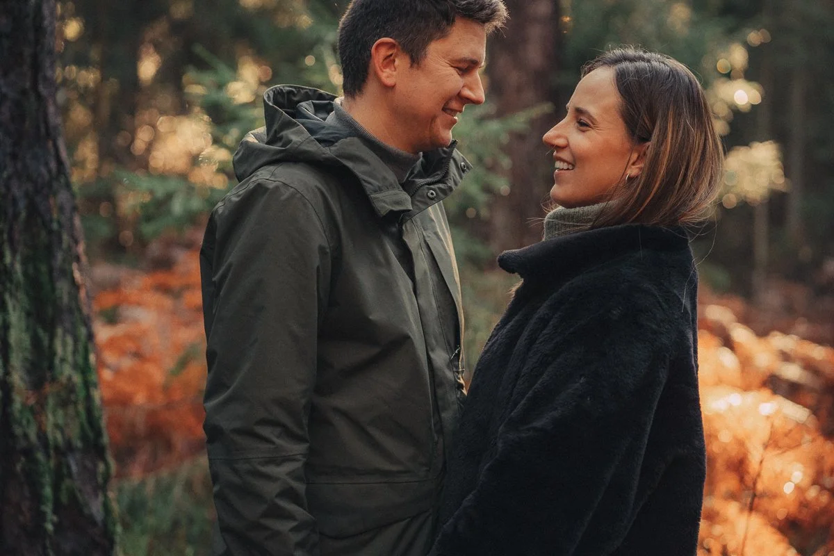 A man and woman smiling and looking into each other's eyes outdoors in a forest, standing close together with trees and fall foliage in the background.
