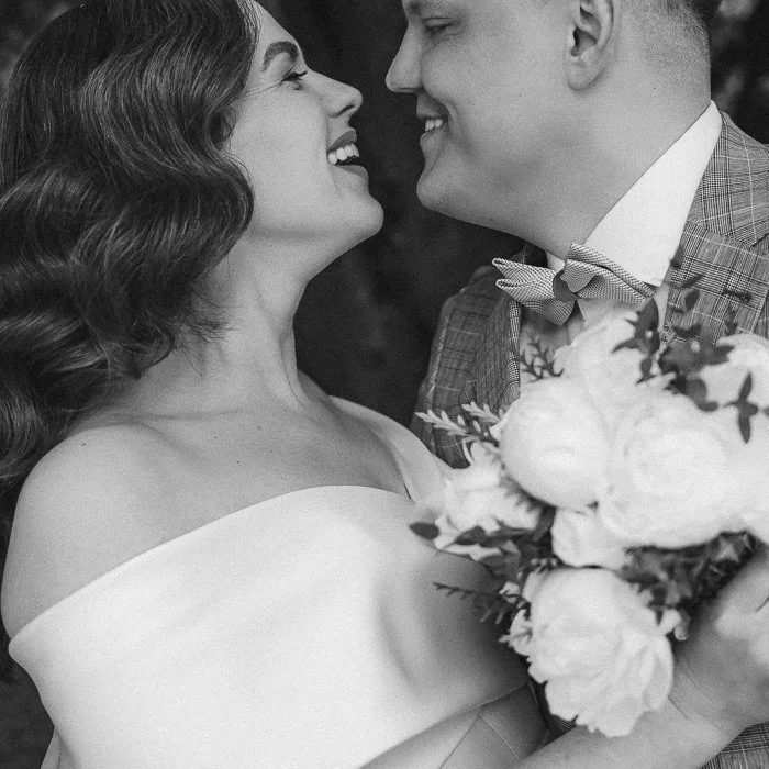 A black and white photo of a bride and groom looking at each other, smiling, with the bride holding a bouquet of flowers.