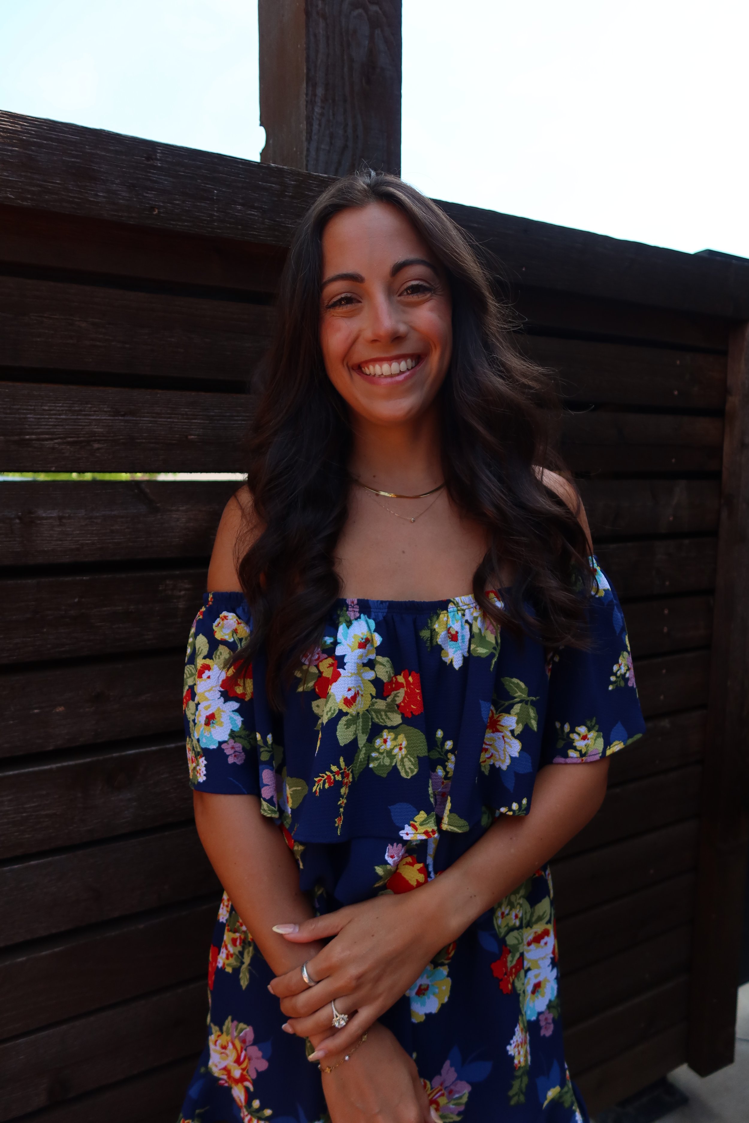 A smiling woman with long dark hair standing against a wooden fence outdoors.