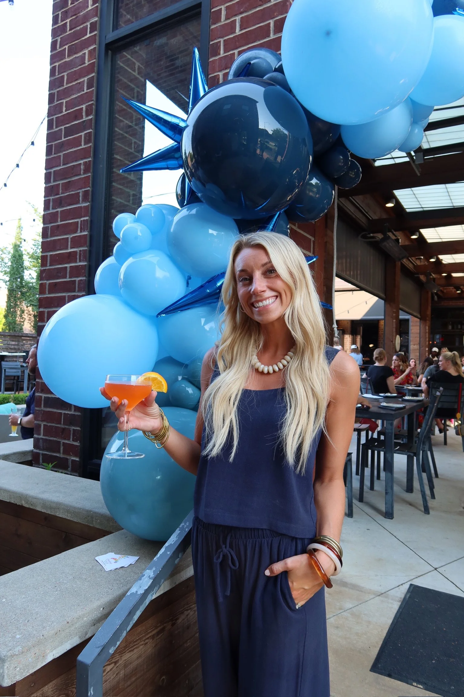 A woman with long blonde hair smiling at a celebration, holding a cocktail with a slice of orange. She is wearing a navy sleeveless top, pearl necklace, and bracelets. Behind her, there are blue and black balloons and a brick building with large windows, with people gathered at a social event outdoors.