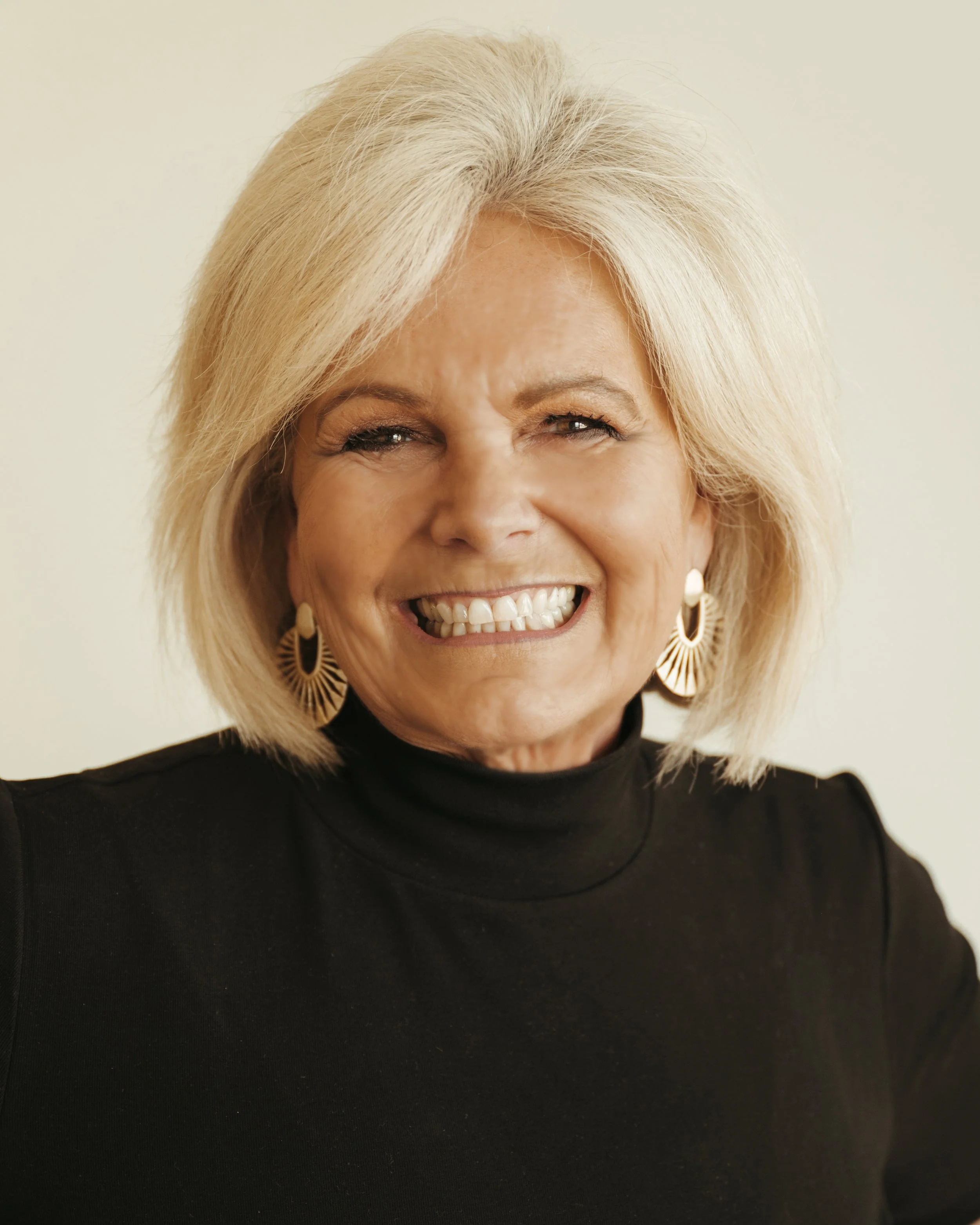 A smiling elderly woman with short, blonde hair, wearing large, gold, sunburst-style earrings and a black turtleneck top.