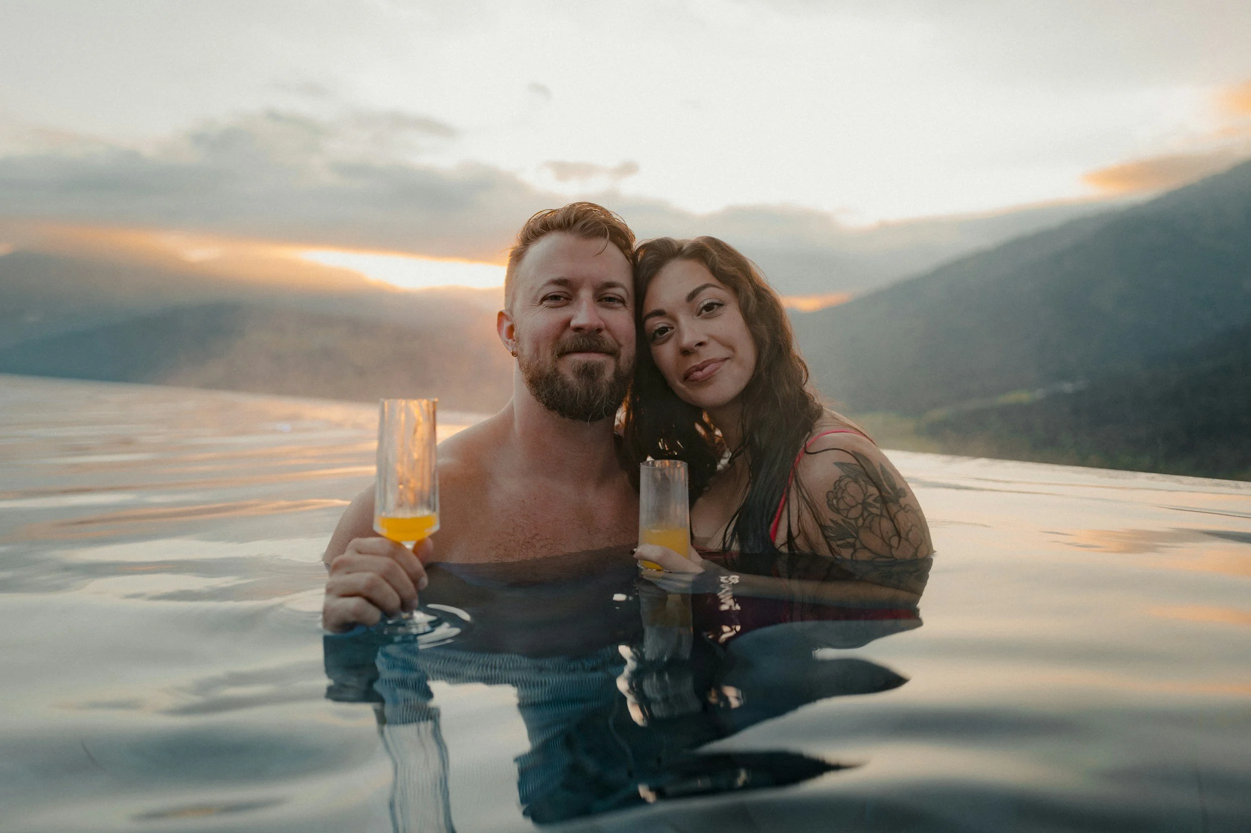A man and woman in a hot tub outdoors at sunset, holding glasses of orange drinks, smiling at the camera.