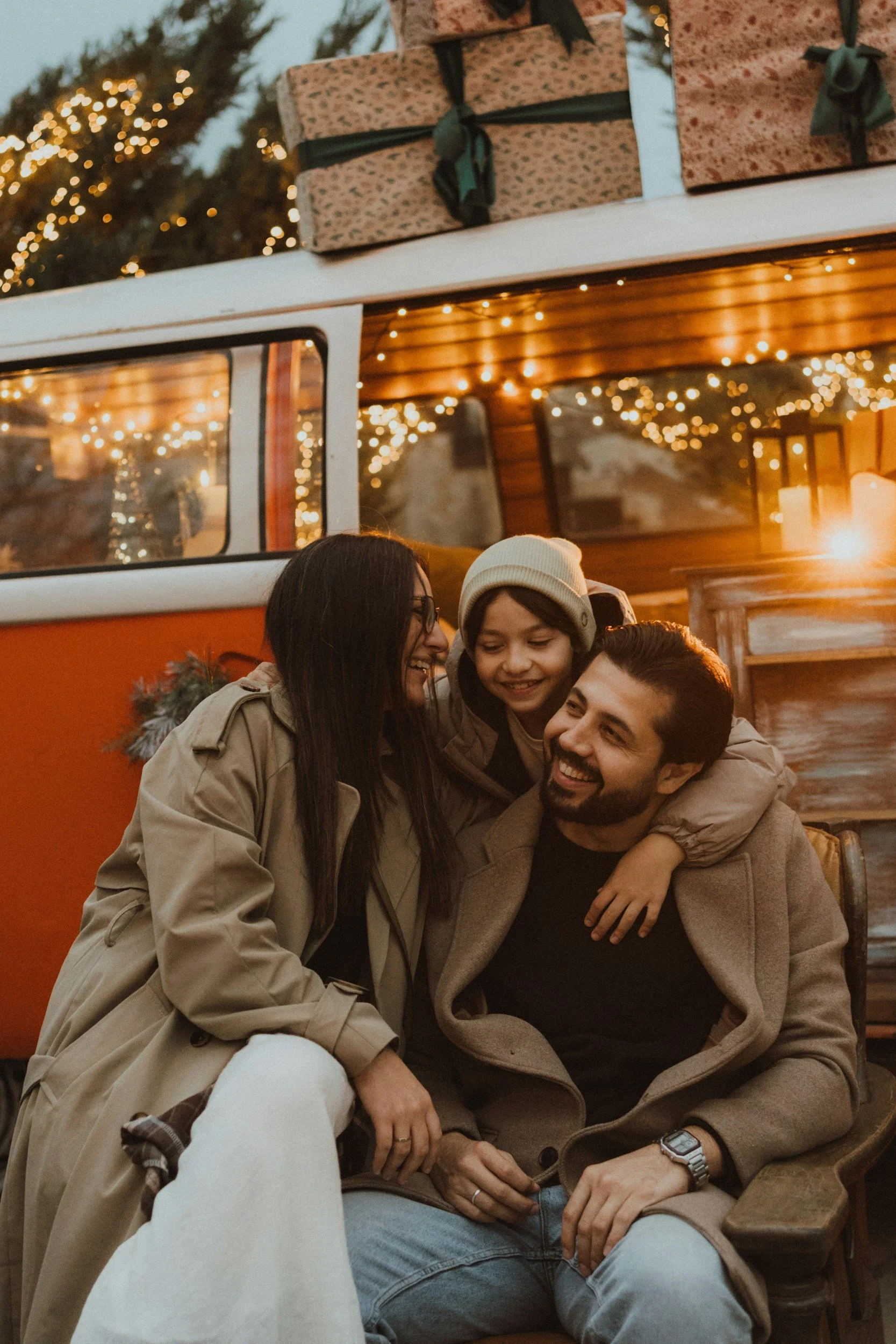 A group of four friends, two men and two women, sitting together outside near a wooden bench, smiling and hugging, during a festive evening with string lights and decorated Christmas trees in the background. They are dressed warmly.