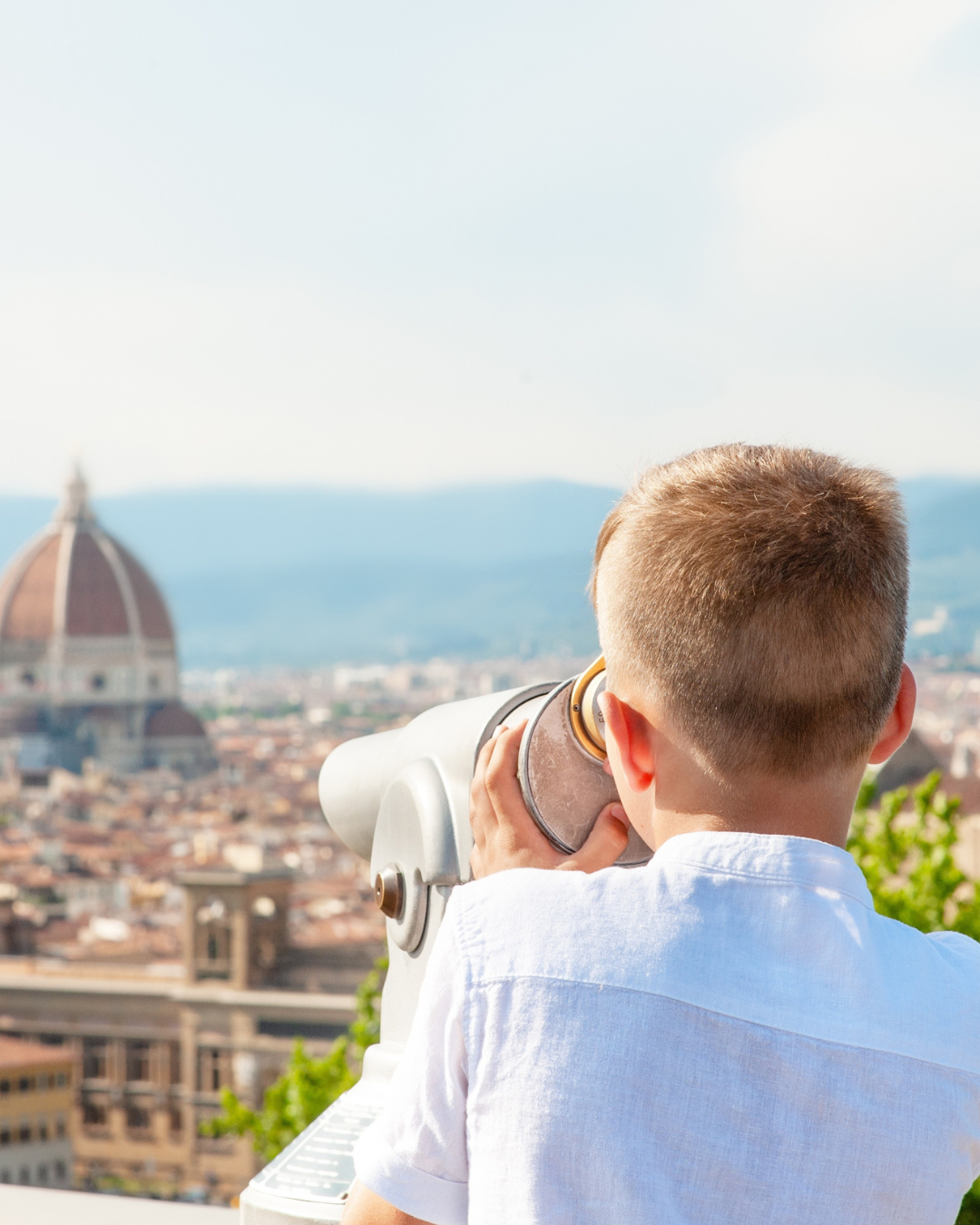 A young boy with short hair looking through a coin-operated telescope at the cityscape of Florence, Italy, with the Florence Cathedral's dome in the background.