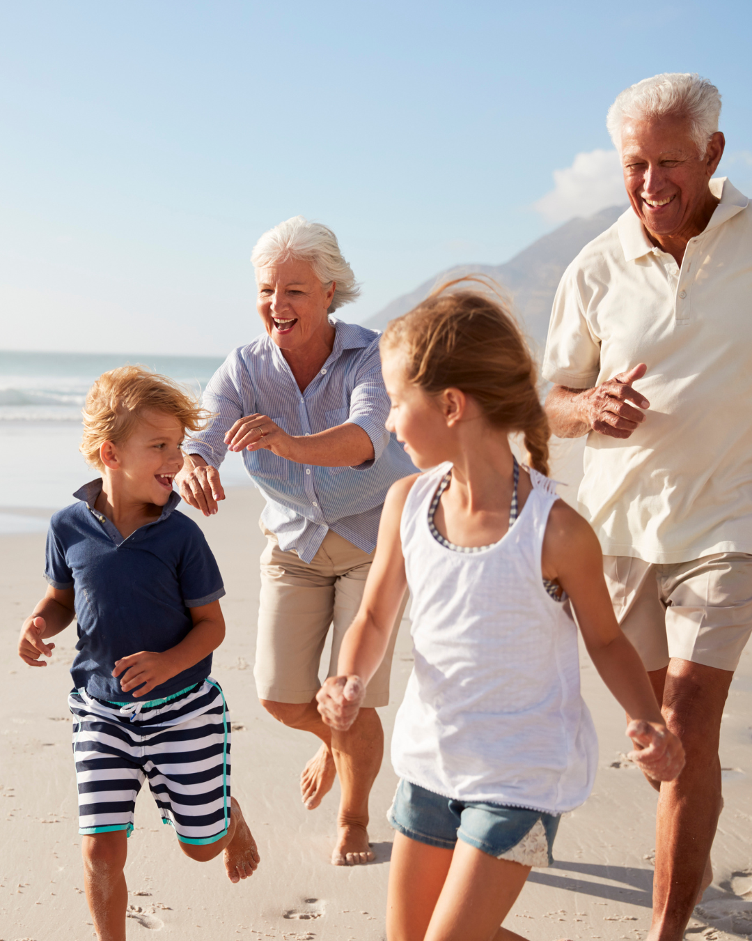 A multigenerational family of two older adults and two children running and playing on a sunny beach with mountains in the background.