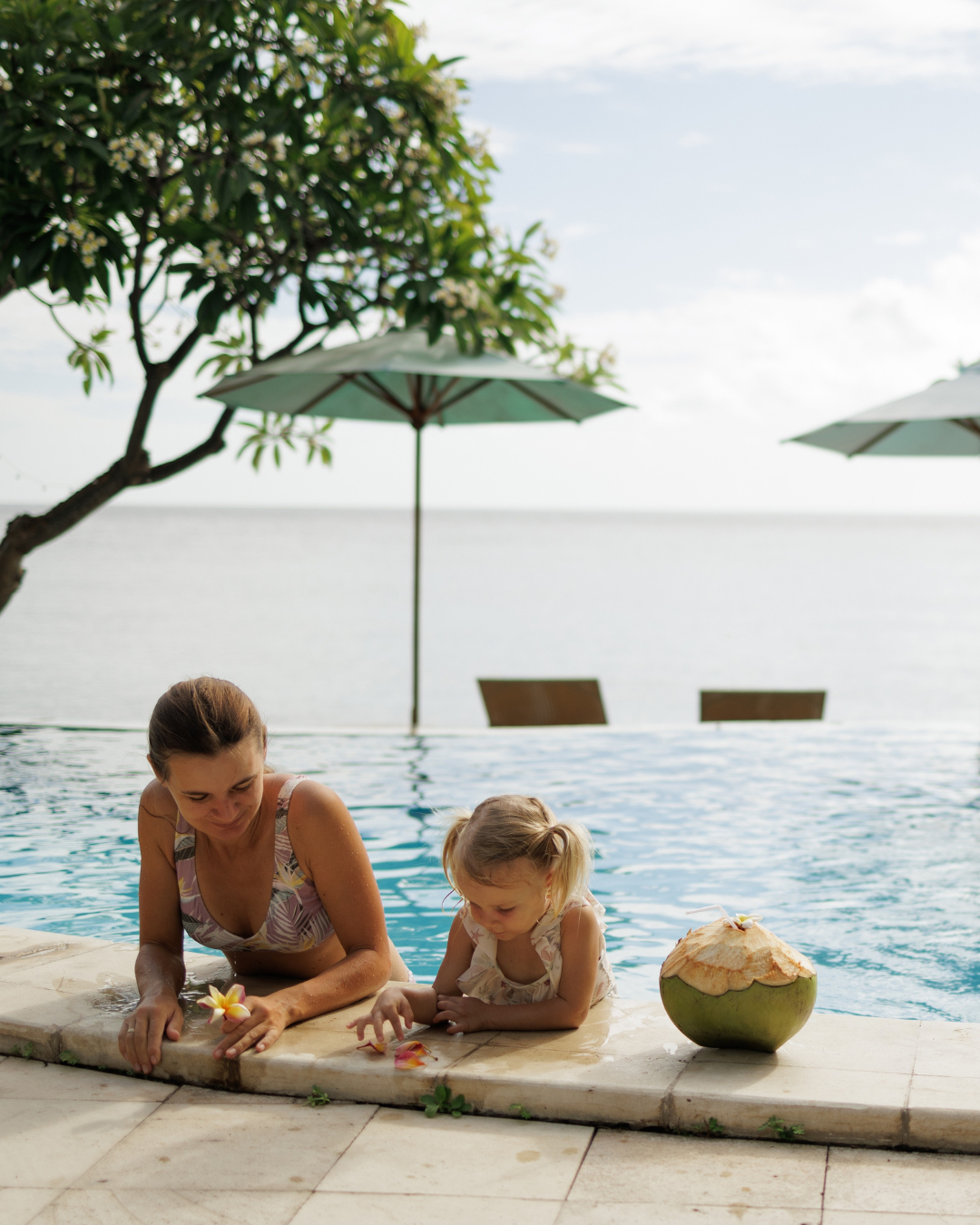 Woman and a young girl relaxing by a swimming pool with a coconut drink, umbrellas, and a tree in the background.