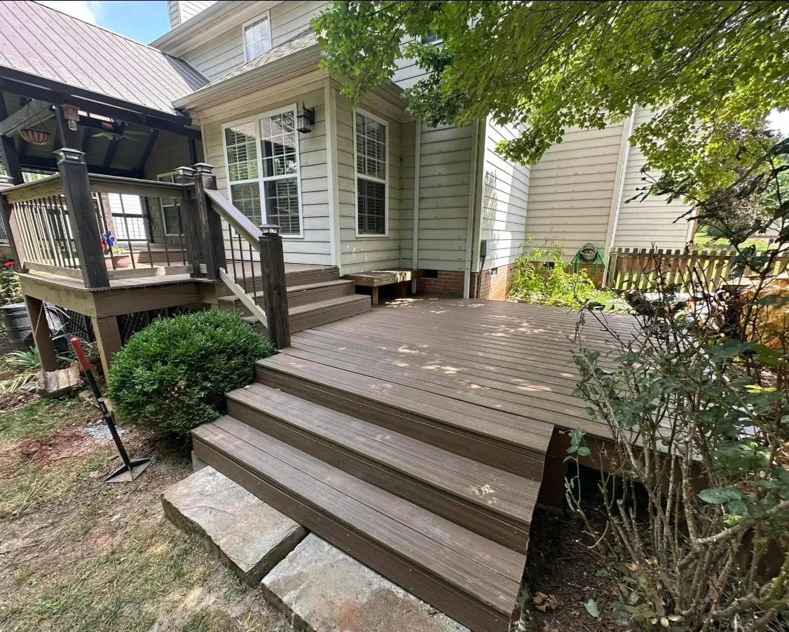 Photo of a backyard deck attached to a house, with wooden steps leading down to the yard, surrounded by greenery.