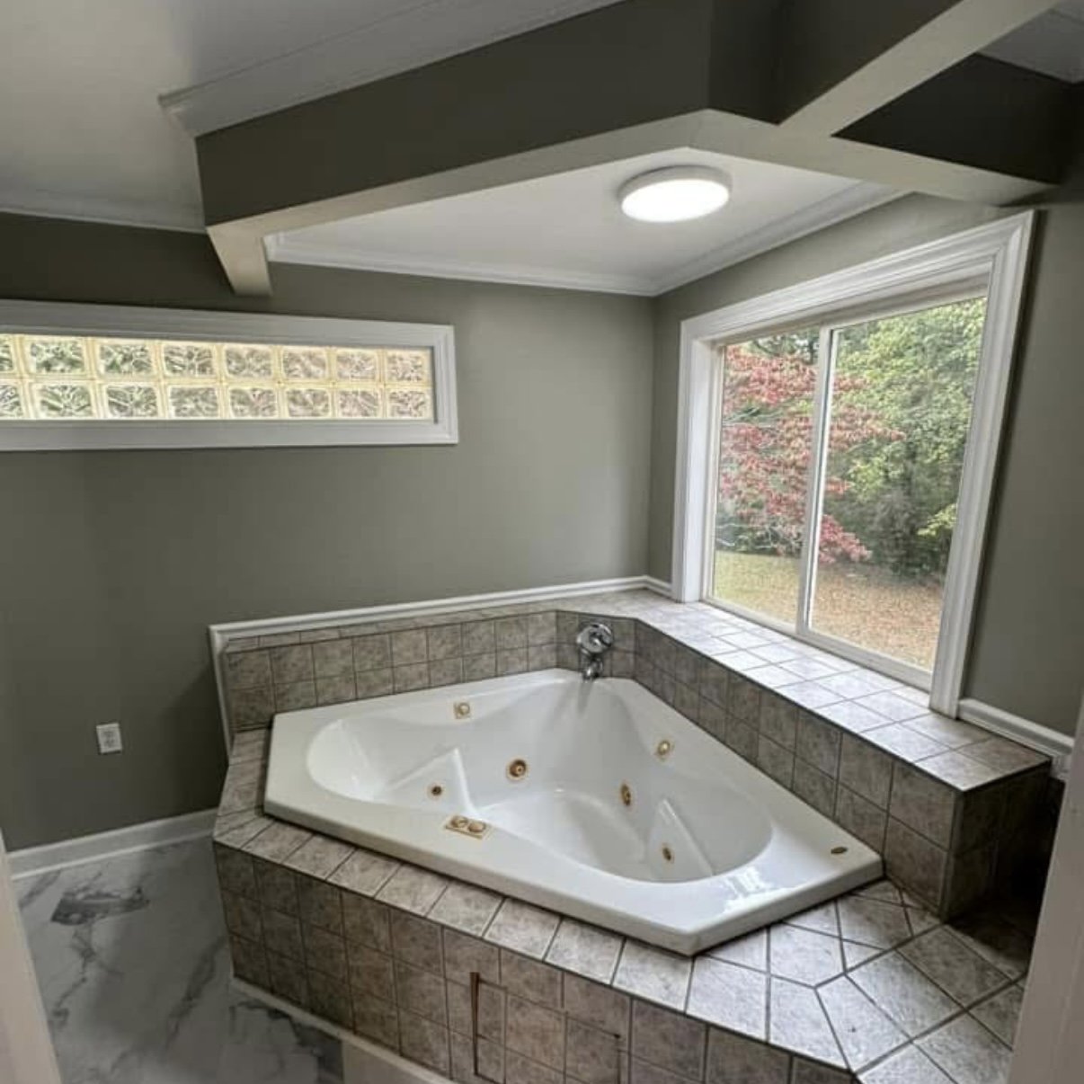 Bathroom with large corner bathtub surrounded by brown tiles, a window with view of a garden with trees, a small horizontal window, and a ceiling light.