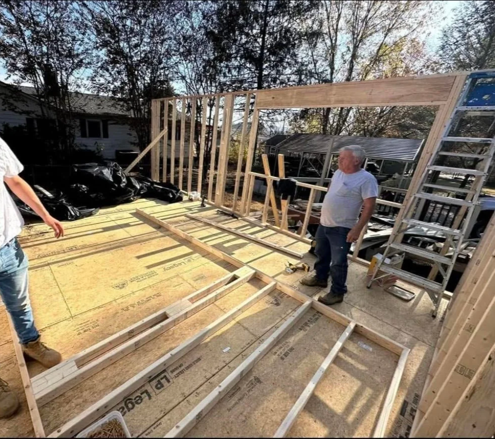 Two men building a wooden frame for a house in the backyard, with construction tools and ladders visible.