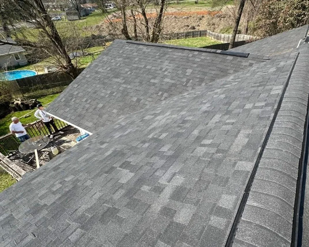 An aerial view of a gray shingle roof with two men standing on a small backyard patio talking, surrounded by trees and a fenced yard.