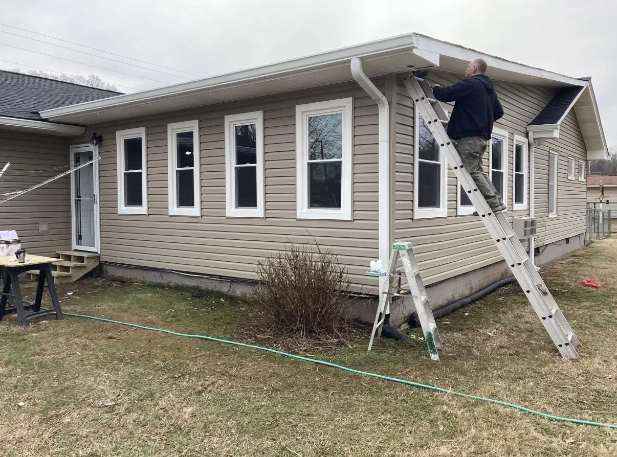 A man standing on a ladder cleaning the gutter of a beige house with white-trimmed windows and a black roof, during daytime.