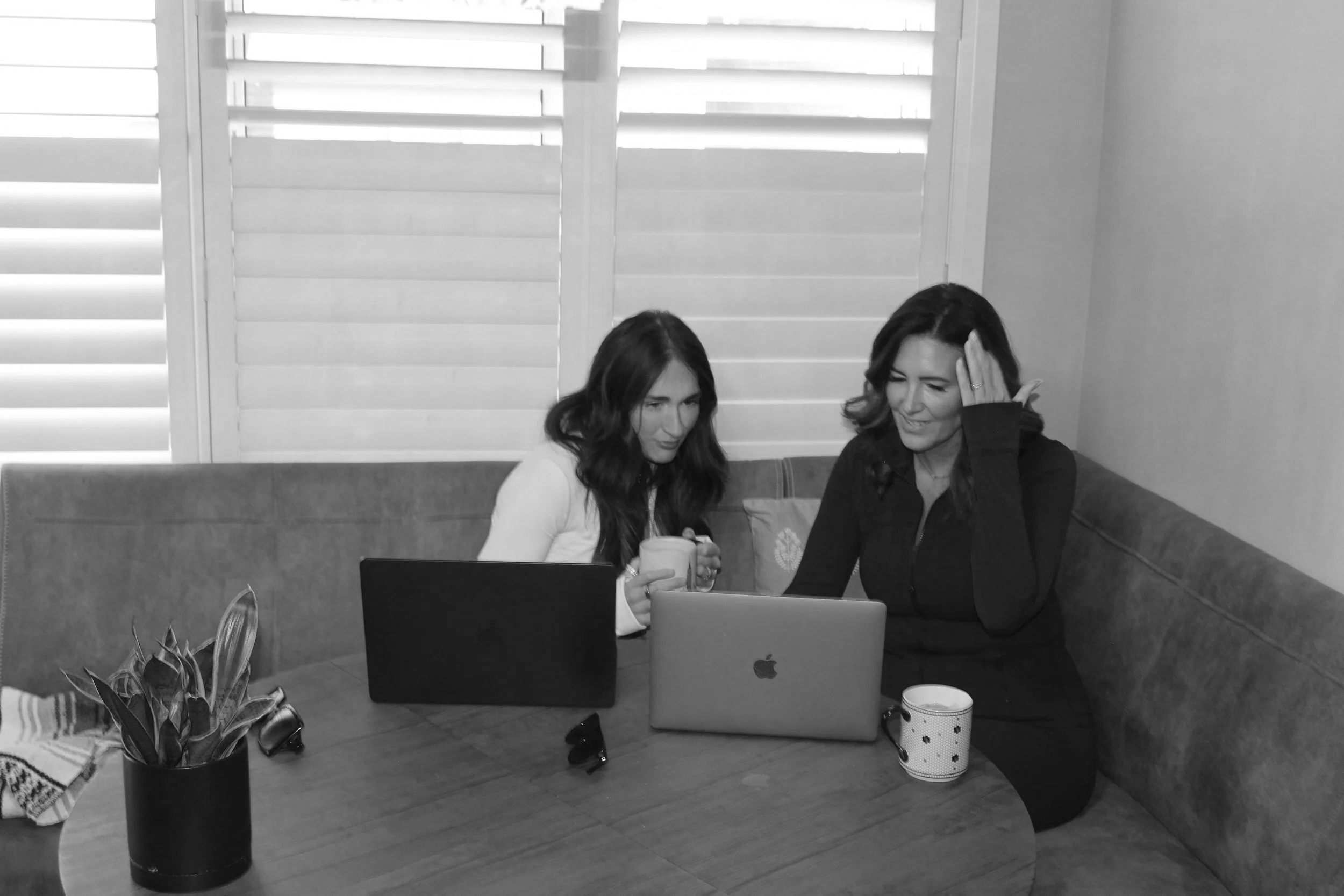 Two women sitting at a round table with laptops and coffee mugs, near window with shutters, engaging in conversation.