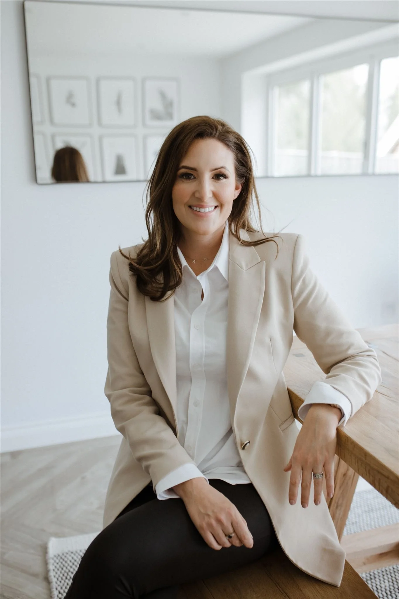A woman with brown hair smiling, wearing a beige blazer and white shirt, sitting at a wooden table in a bright room.