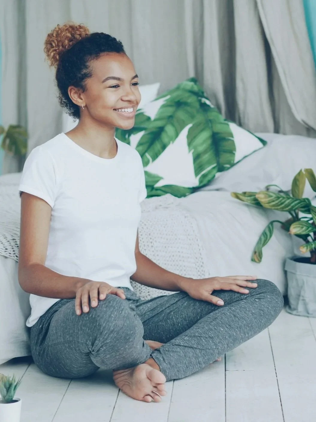 Smile of a woman sitting in a cross-legged yoga pose on the floor in a bedroom, surrounded by tropical plants and leaf-patterned pillows.
