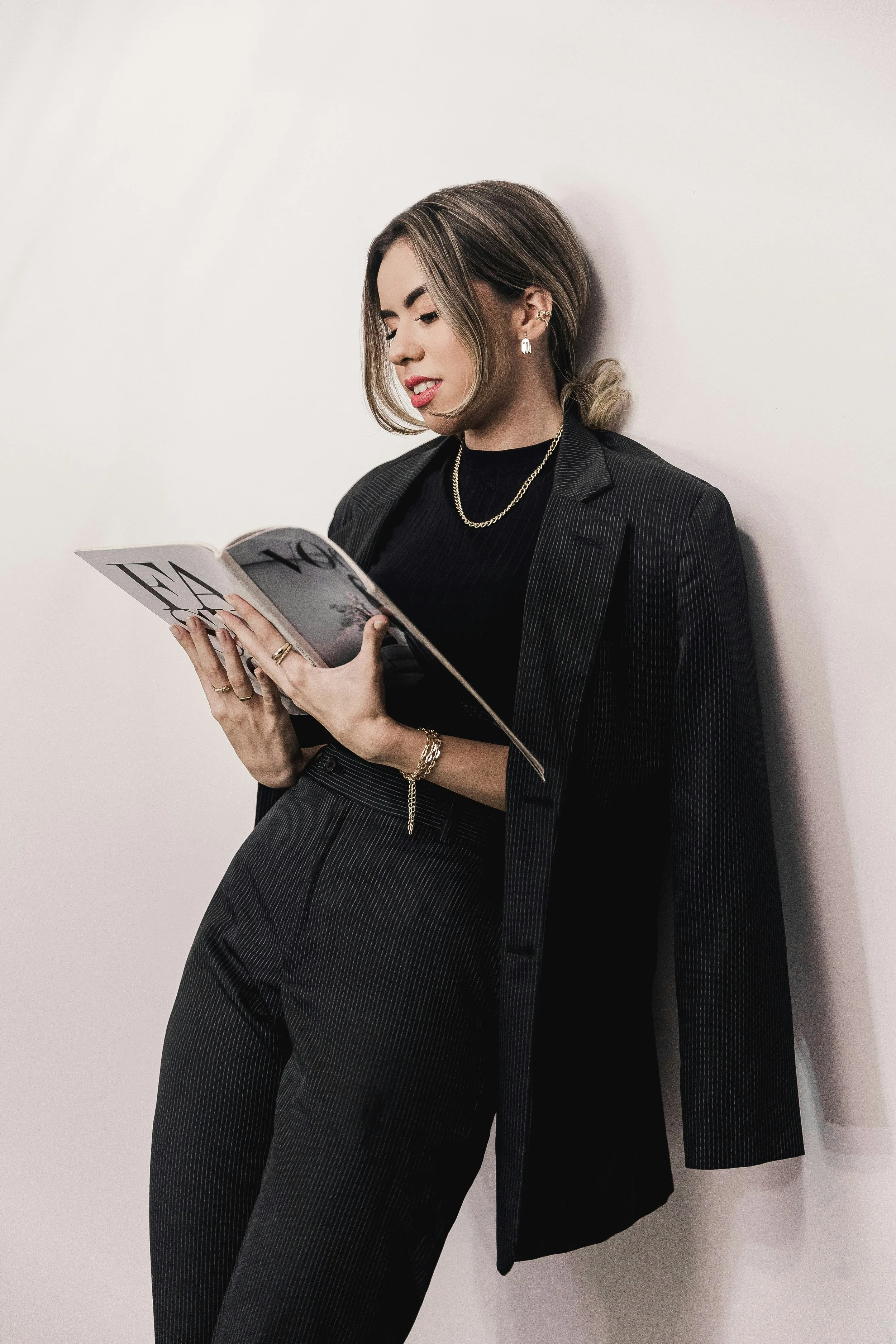 Woman in black pinstripe suit reading a magazine while leaning against a white wall.