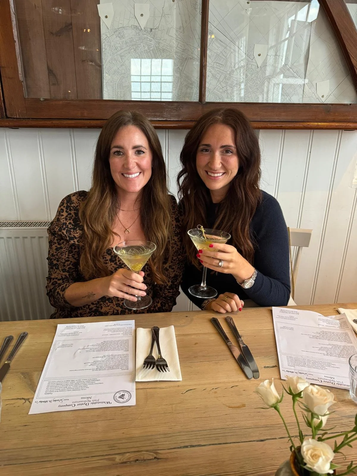 Two women smiling and holding cocktails at a restaurant table, with menus, utensils, and a flower arrangement in the foreground.