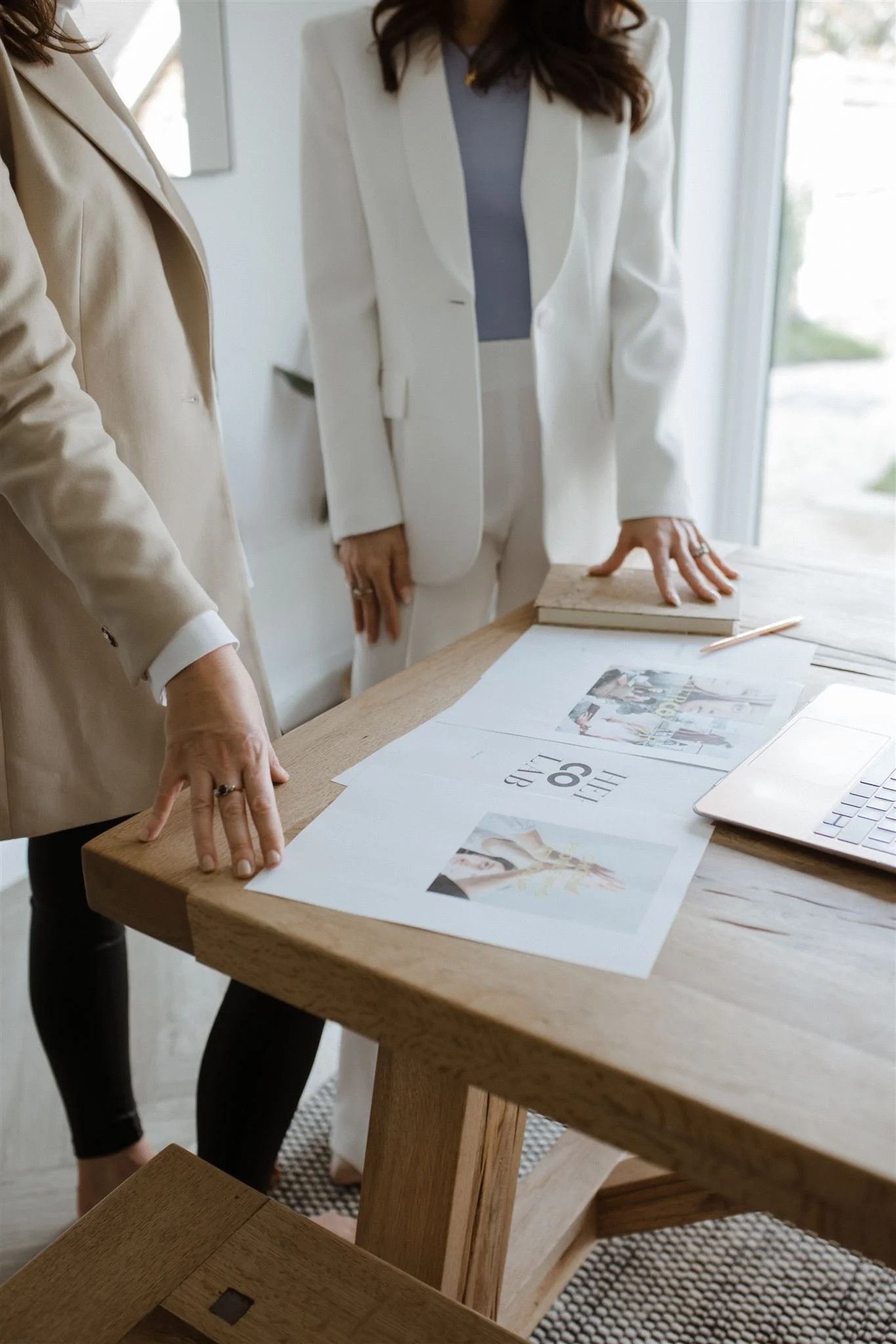 Two women in white blazers standing at a wooden table with papers, photos, a notebook, and a laptop on it, discussing a project in a bright room with natural light.