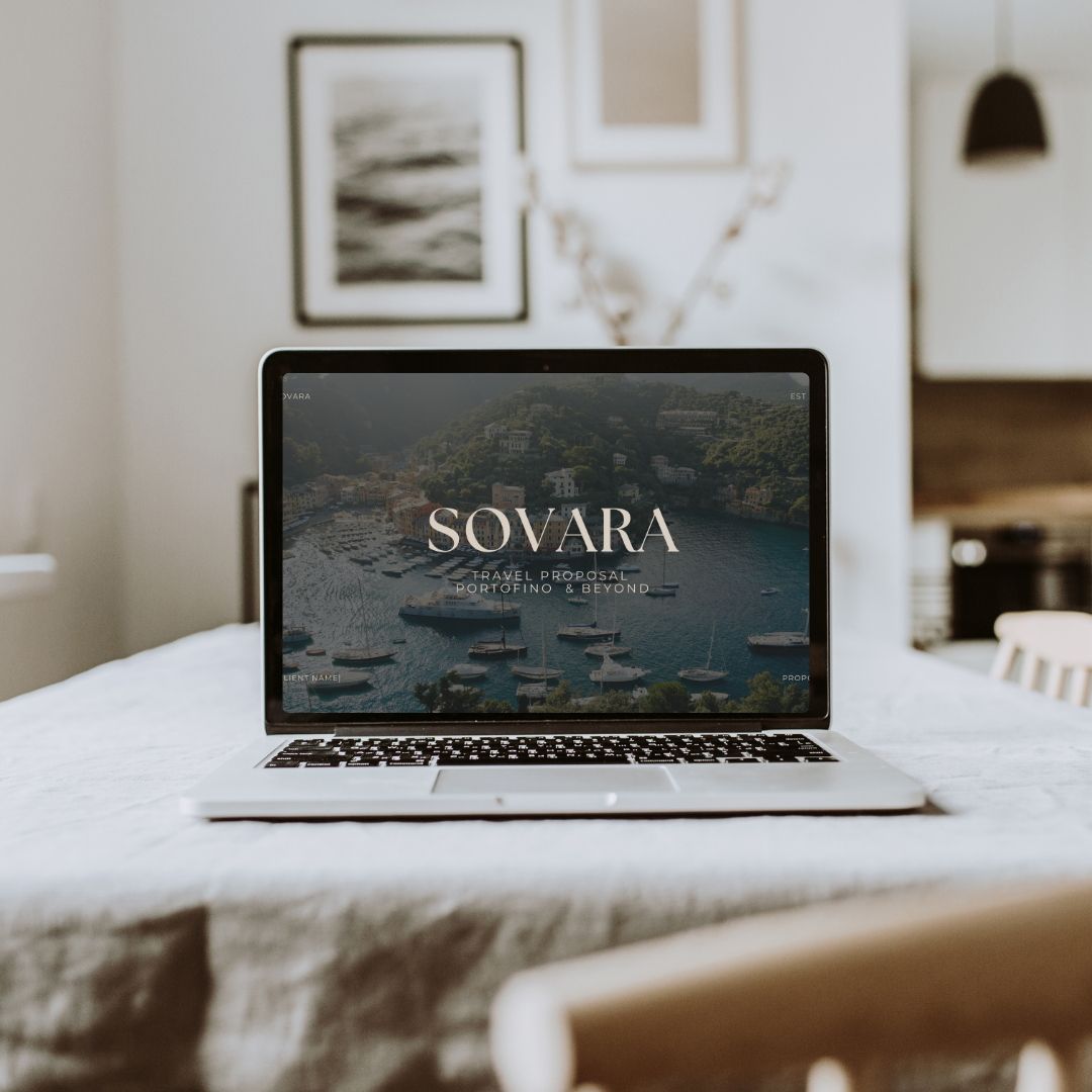 Open laptop on a dining table displaying a travel proposal website titled "Sovara" with a waterfront scene of boats and buildings in a hilltop town, in a bright, modern dining room with wall art and a black pendant lamp.