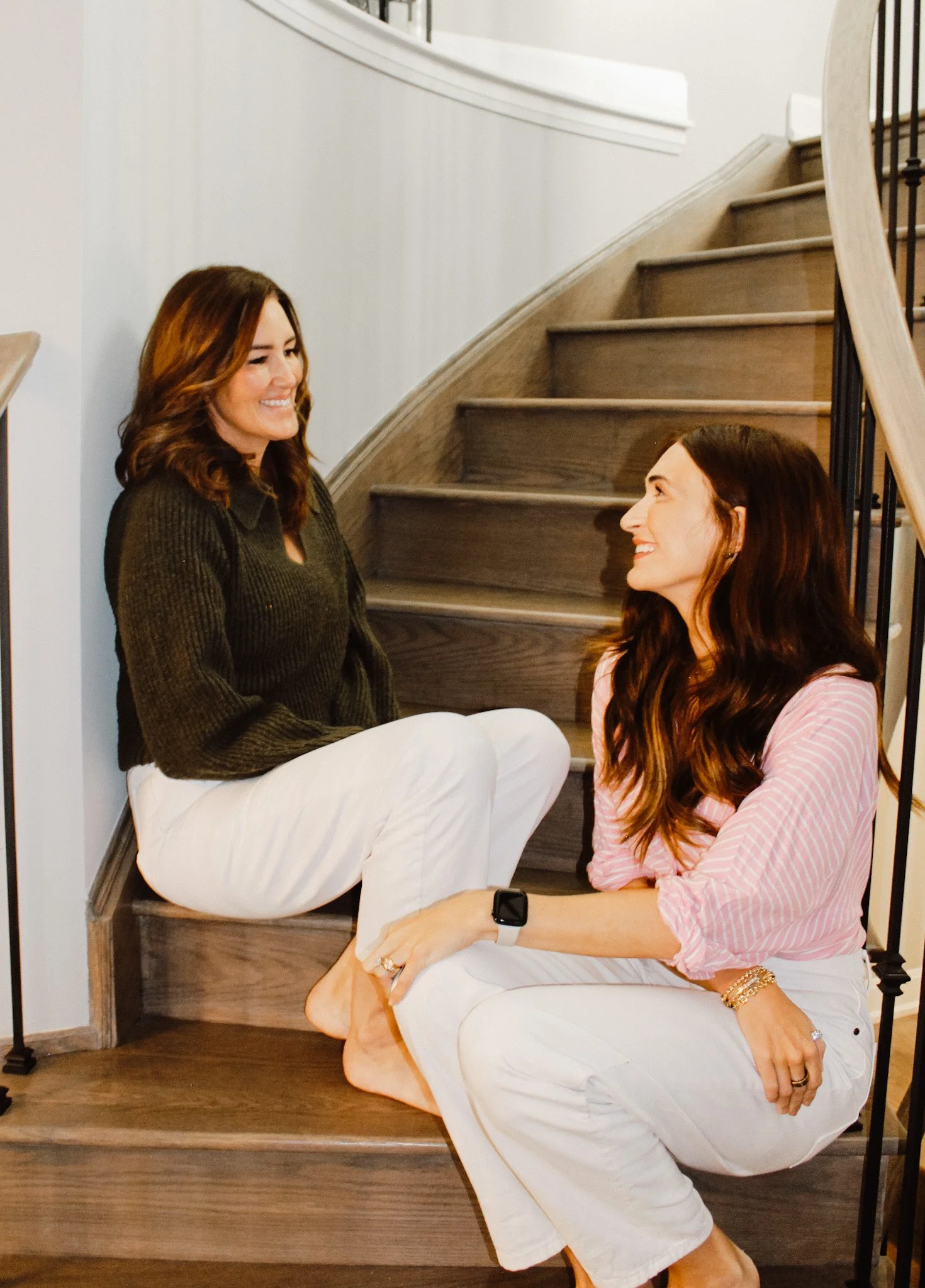 Two women sitting on wooden stairs, smiling at each other, with one woman sitting on the floor and the other sitting on the stairs.