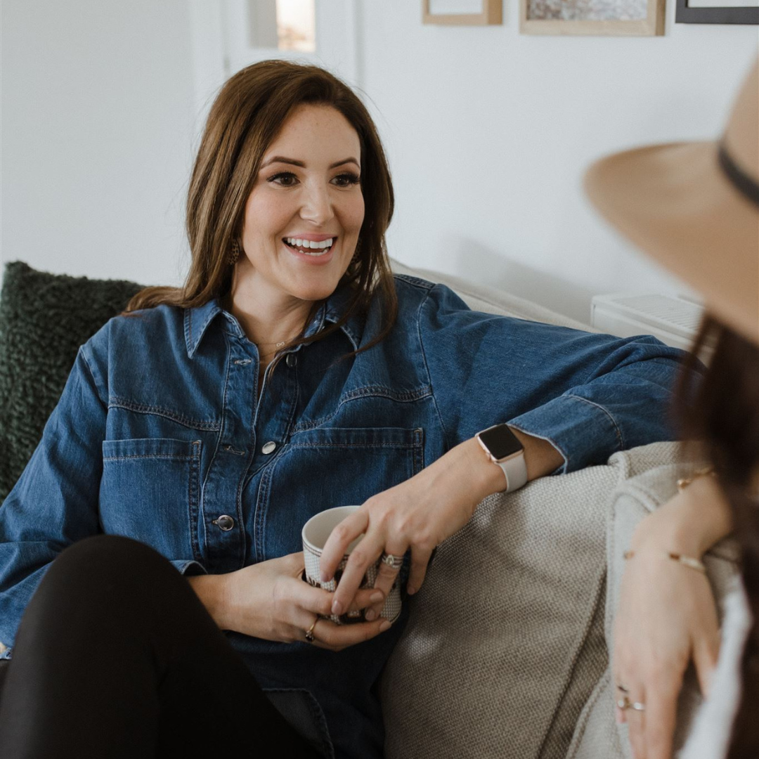 Two women sitting on a sofa, engaged in conversation, one holding a mug, in a cozy living room.