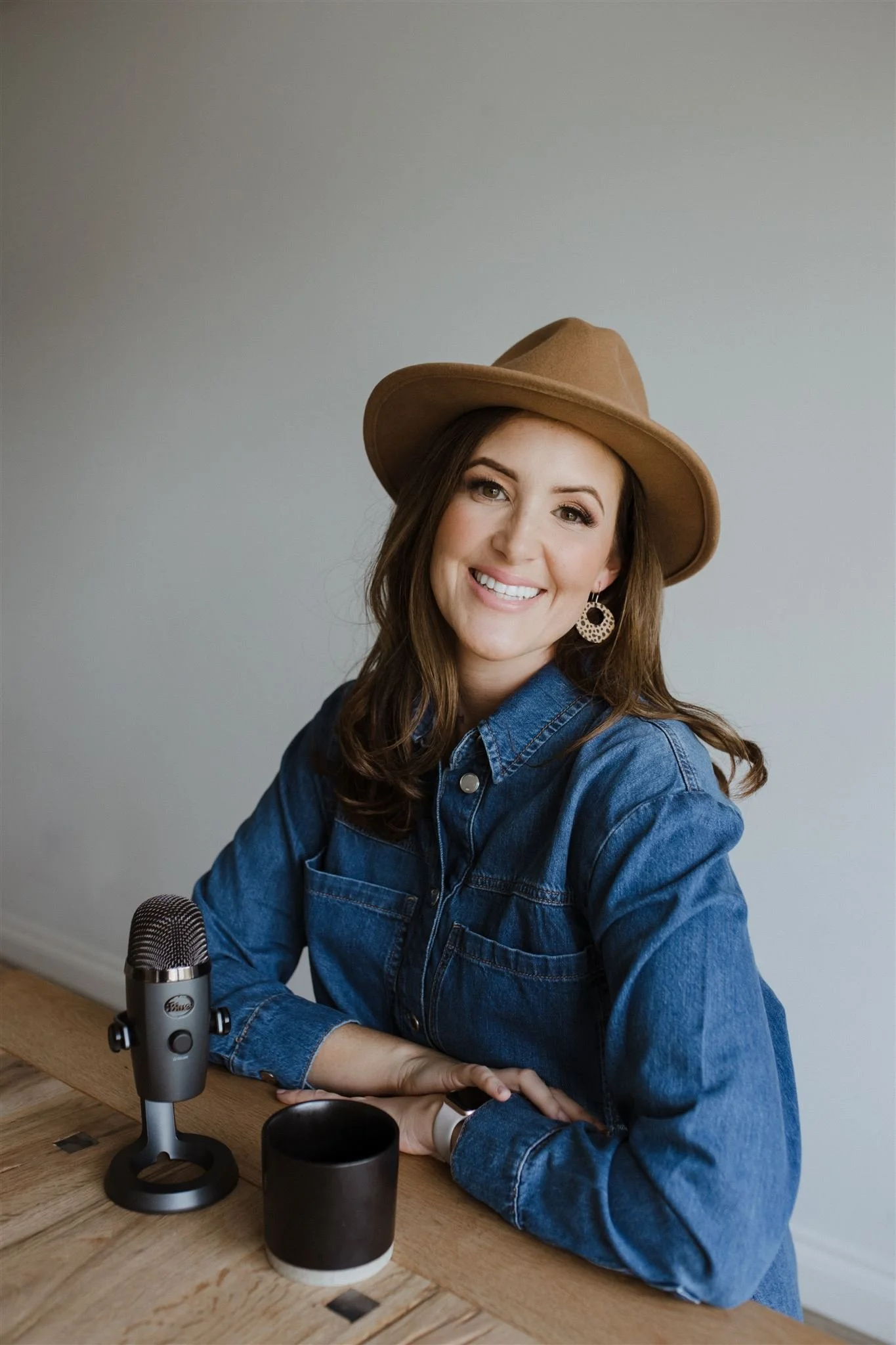 A woman sitting at a wooden table with a microphone and a black mug in front of her. She is smiling, wearing a brown hat, denim shirt, and earrings, with long brown hair against a plain wall background.