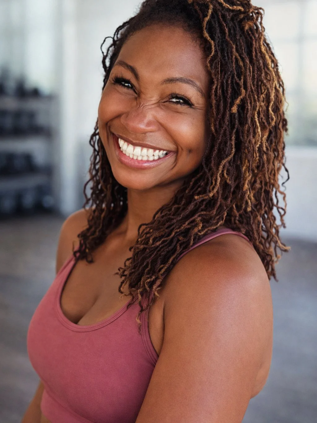 Smiling woman with long, curly hair wearing a pink sports top indoors.