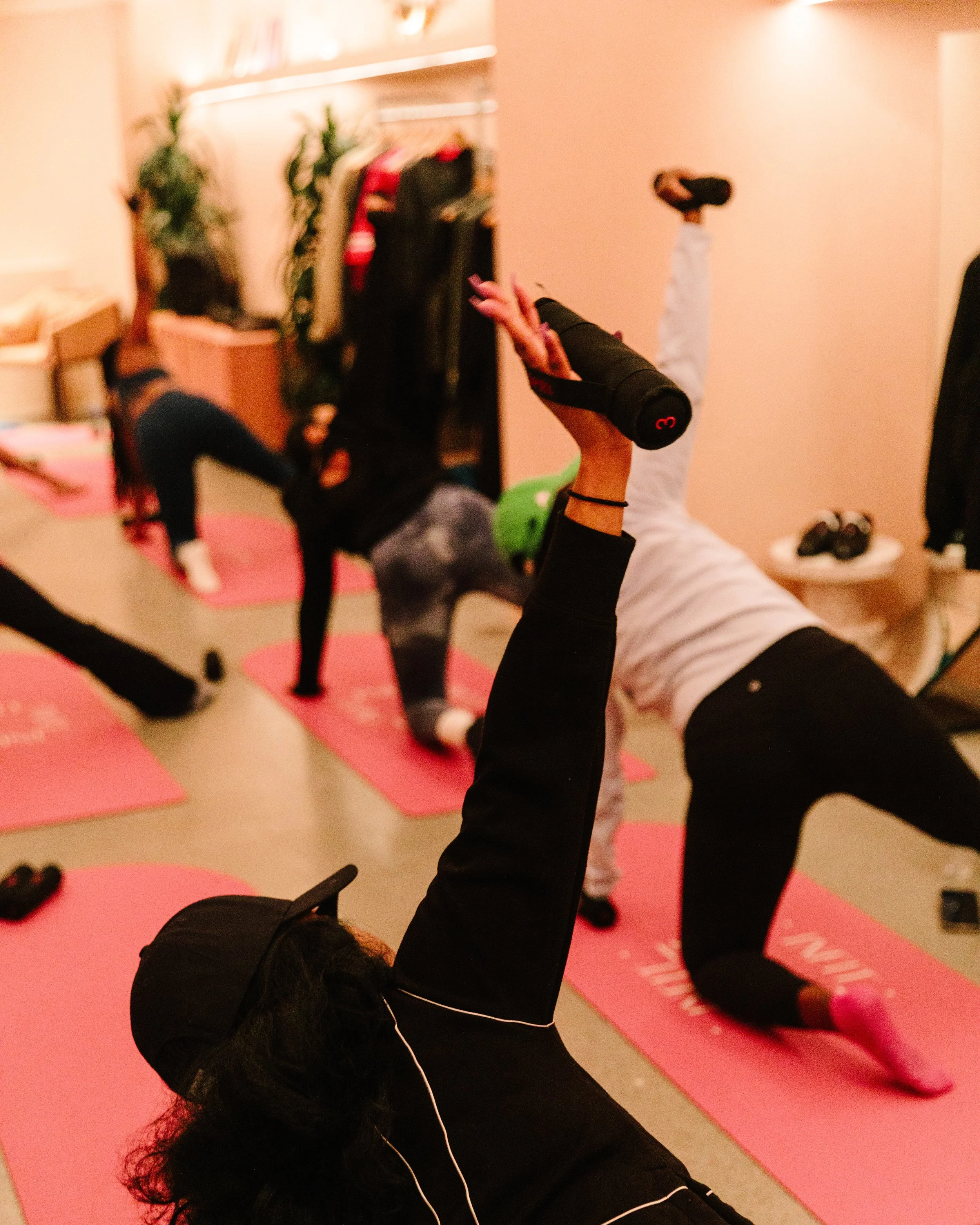 A person lying on a pink yoga mat practicing a handstand or downward dog pose in a room with pink walls. Other people are in the background, some seated on pink mats and others standing, with a few plants and a shelf with books visible.