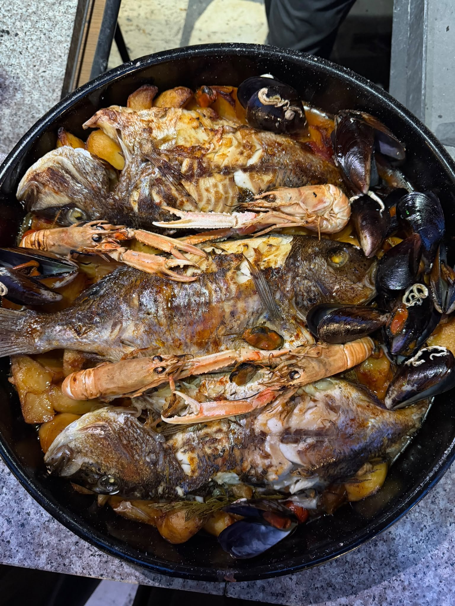 A seafood dish in a black skillet featuring fish, shrimp, mussels, potatoes, and vegetables cooked in a sauce.