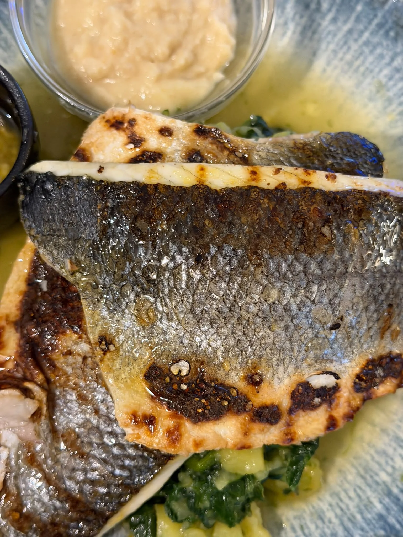 Close-up of a grilled fish fillet with crispy, charred skin and white flesh, resting on a bed of cooked greens and yellow mashed potatoes.
