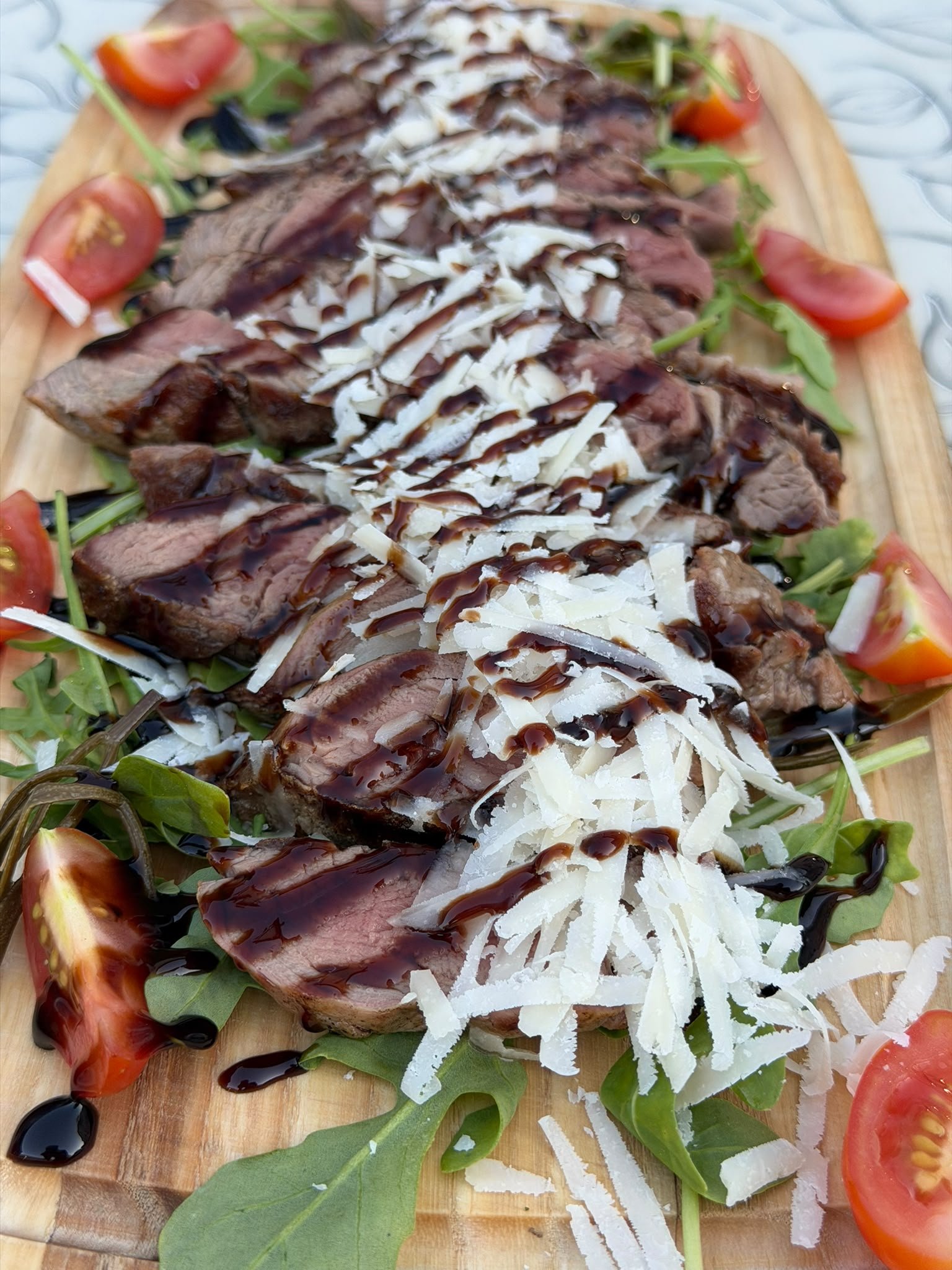 Slices of cooked steak topped with shredded cheese and drizzled with balsamic glaze, garnished with cherry tomato halves and arugula on a wooden board.