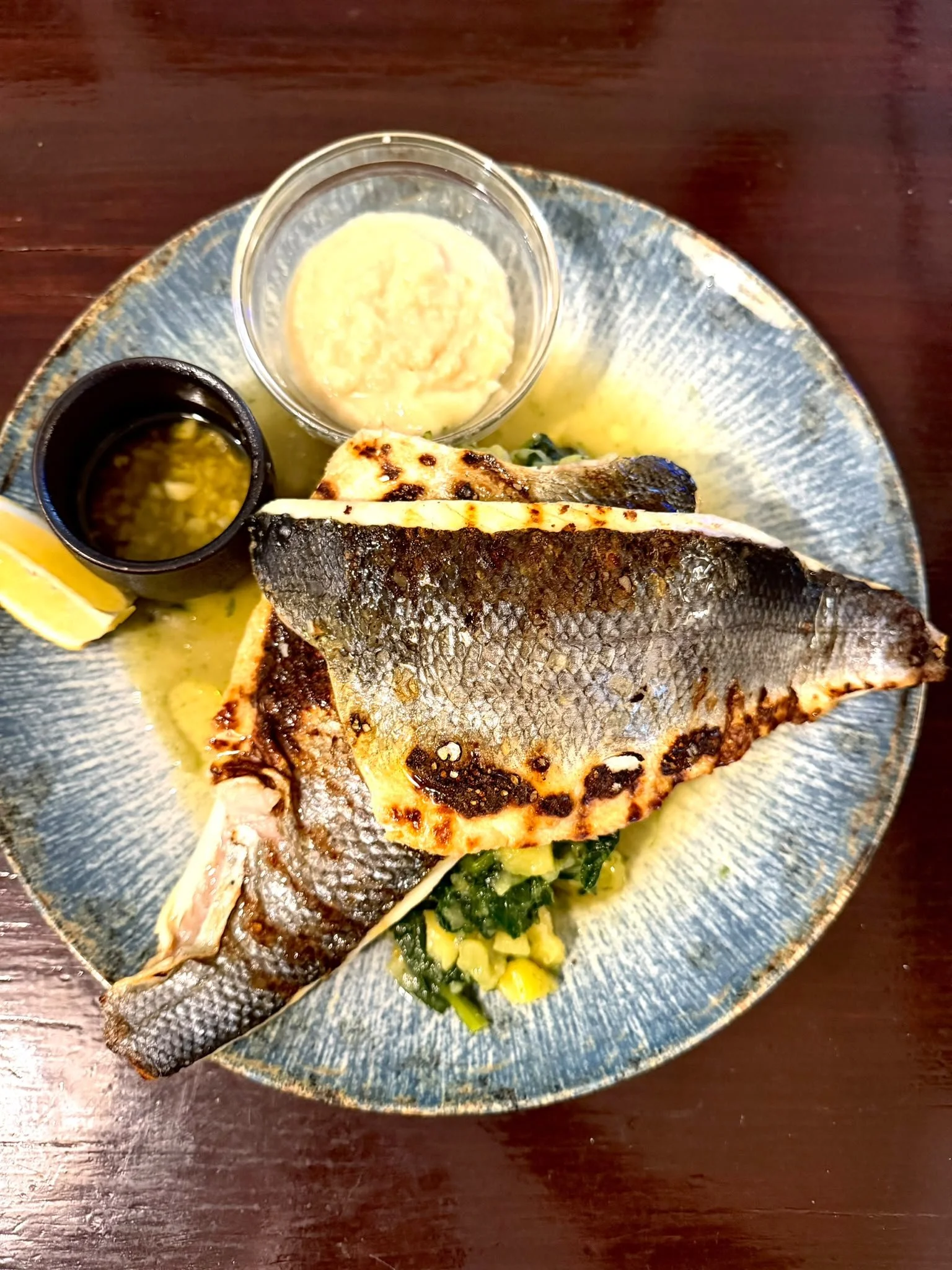 Grilled fish with lemon wedge, roasted garlic, and condiments on a decorative plate.