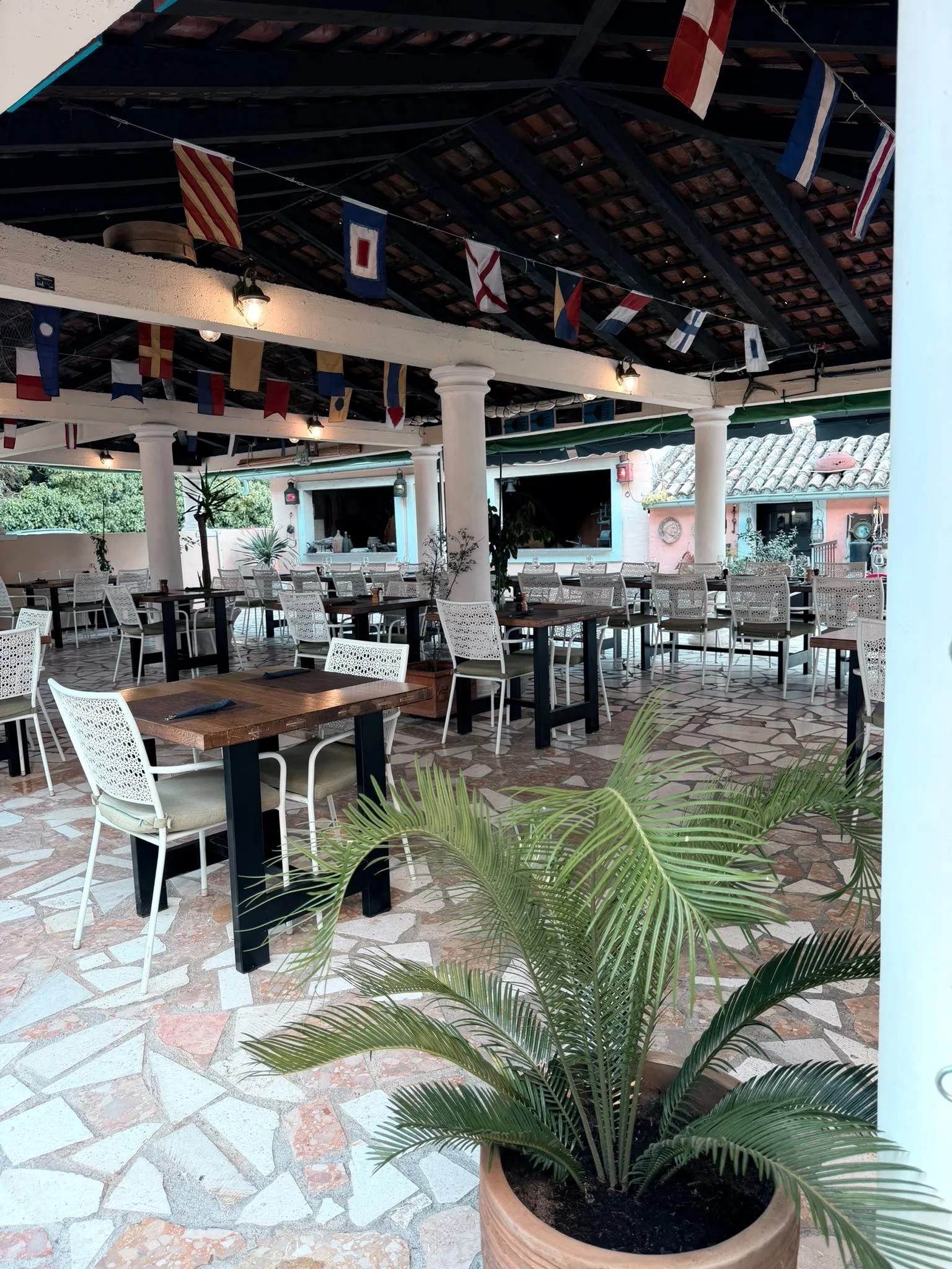 An outdoor dining area with tables, chairs, potted plants, and flags hanging from the ceiling.