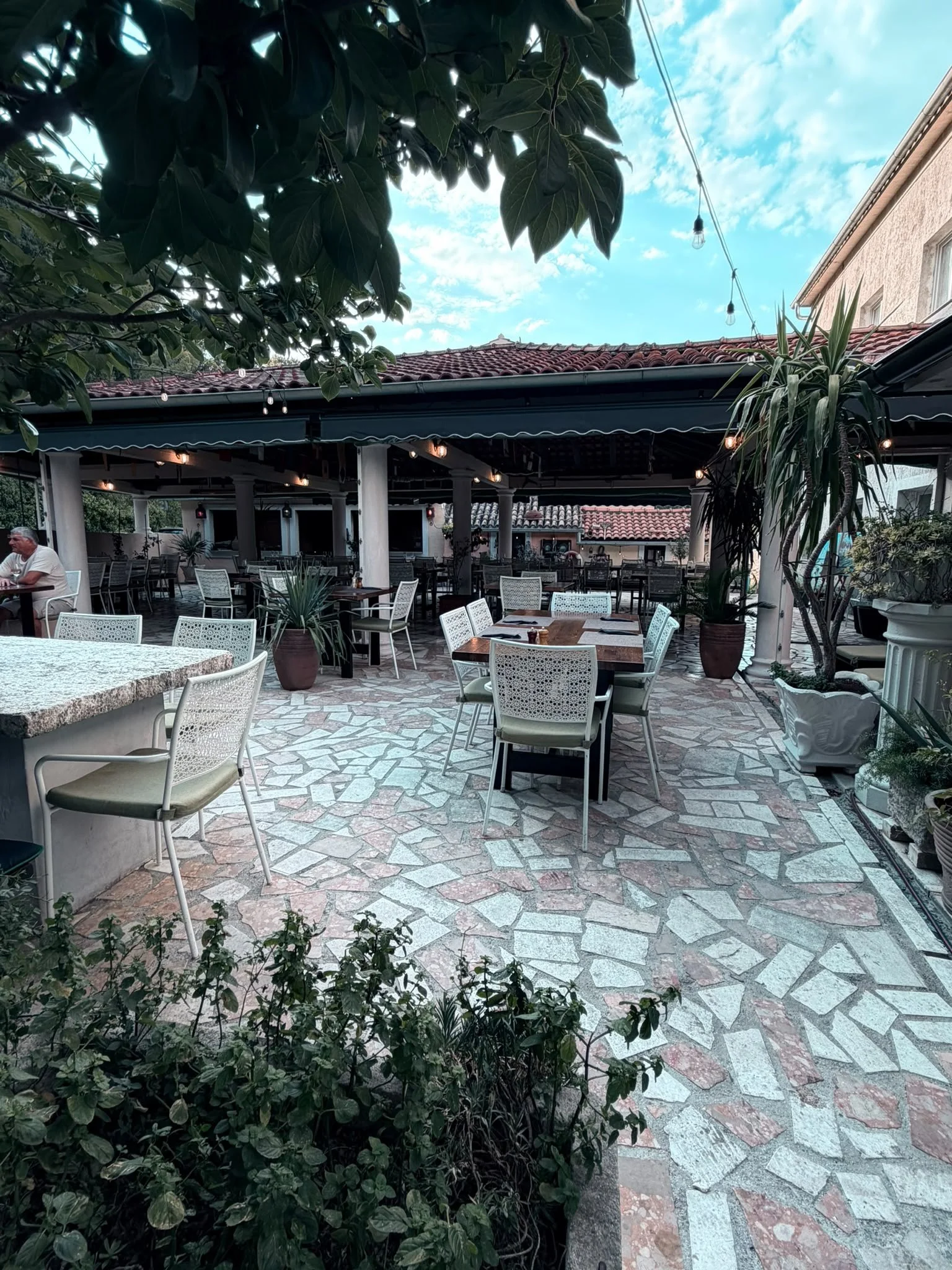 An outdoor patio with white chairs and tables, surrounded by green plants, with a covered roof and string lights overhead, under a partly cloudy sky.