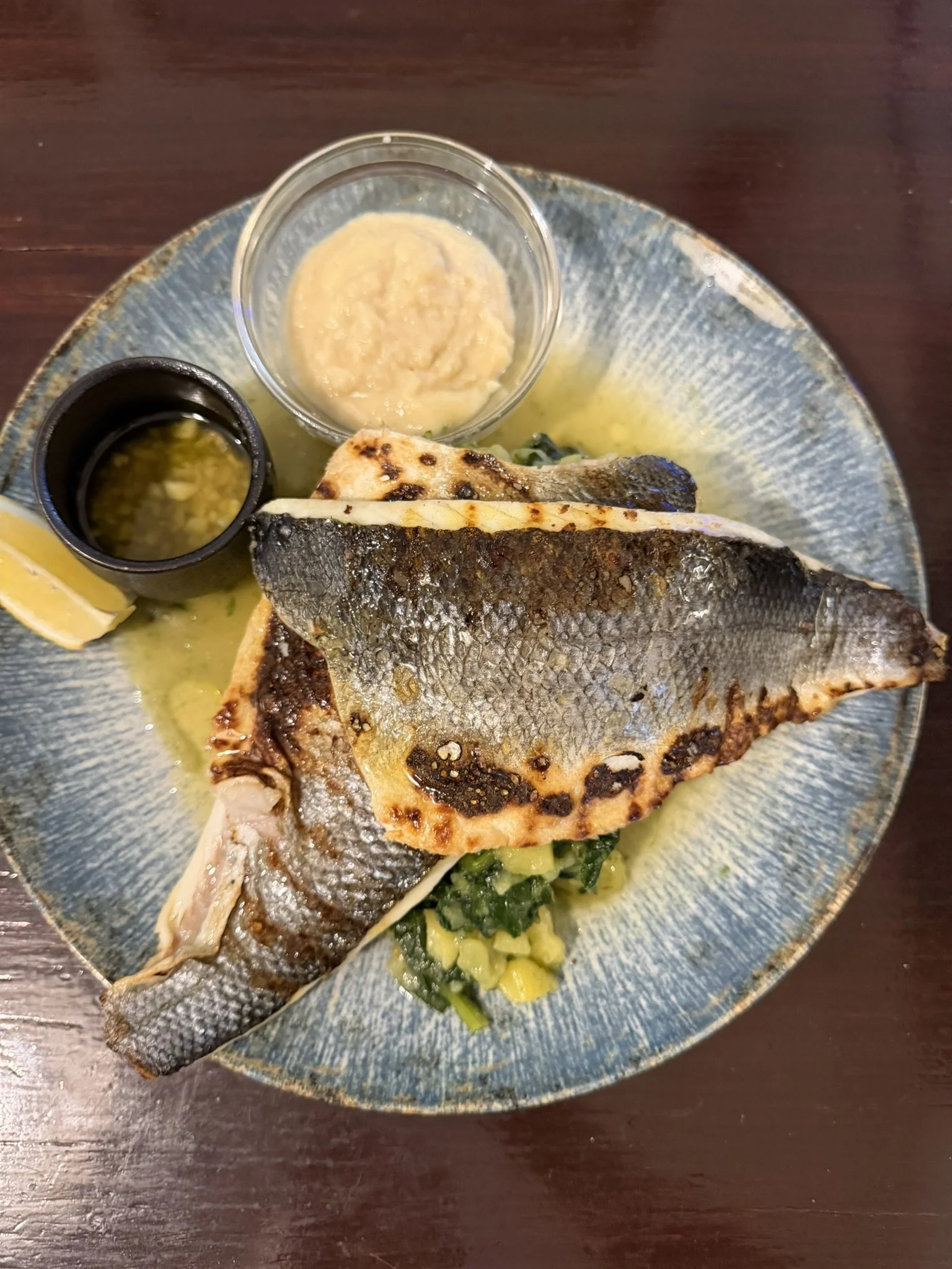 A grilled fish fillet served with lemon, accompanied by a small bowl of sauce, minced garlic, and mashed plantains on a blue ceramic plate.