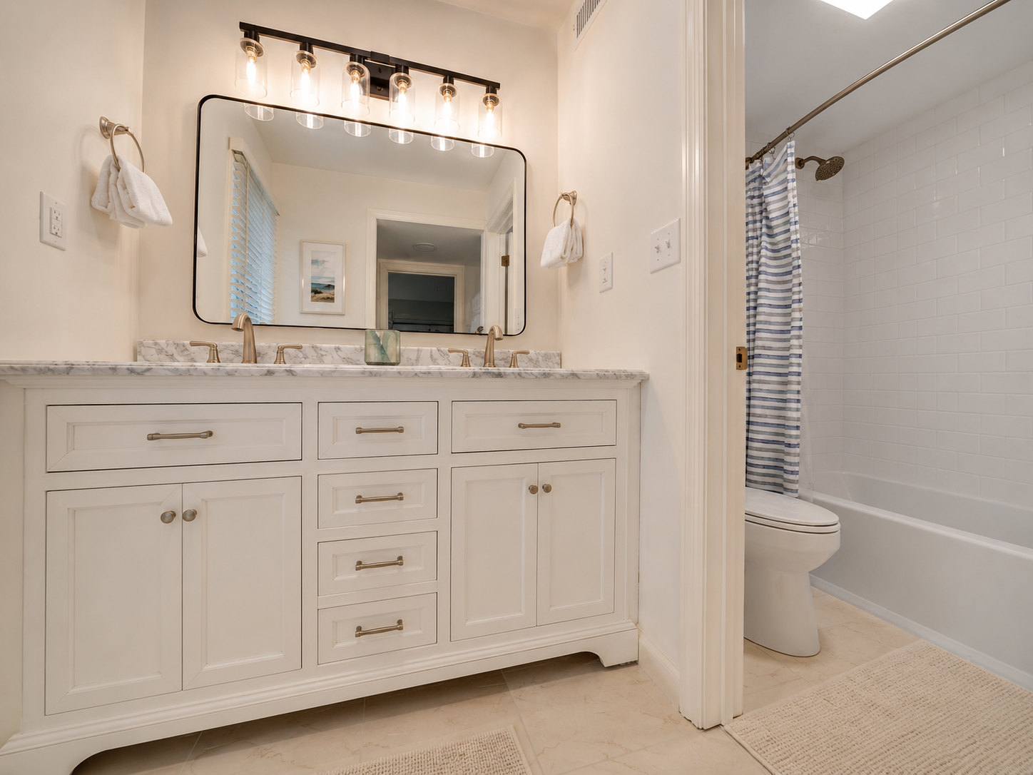 A bathroom with a white vanity with a marble countertop, gold fixtures, and a large mirror. There are two hand towels hanging on the wall, and a window with blinds is reflected in the mirror. To the right, a toilet and a bathtub with a striped shower curtain are visible.