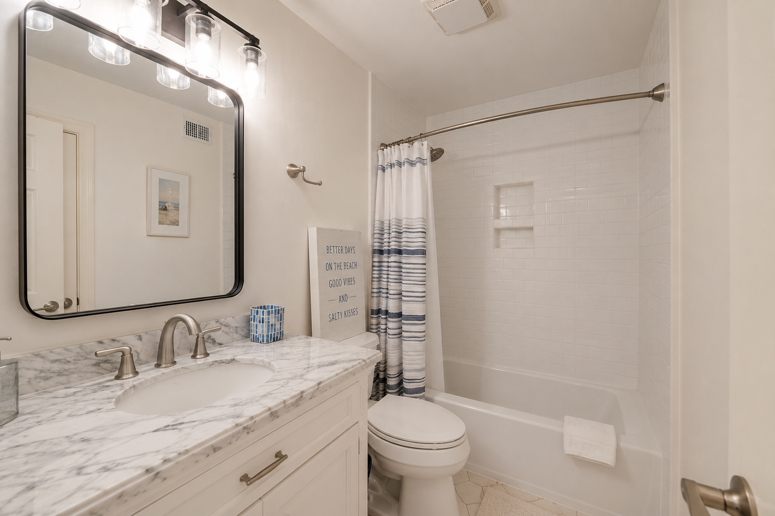 A bathroom with a marble countertop vanity, a mounted mirror, a light fixture above the mirror, a small framed picture, two soap dispensers, a small blue container, a sign on the toilet with beach-themed text, a striped shower curtain, and a bathtub with built-in shelves.