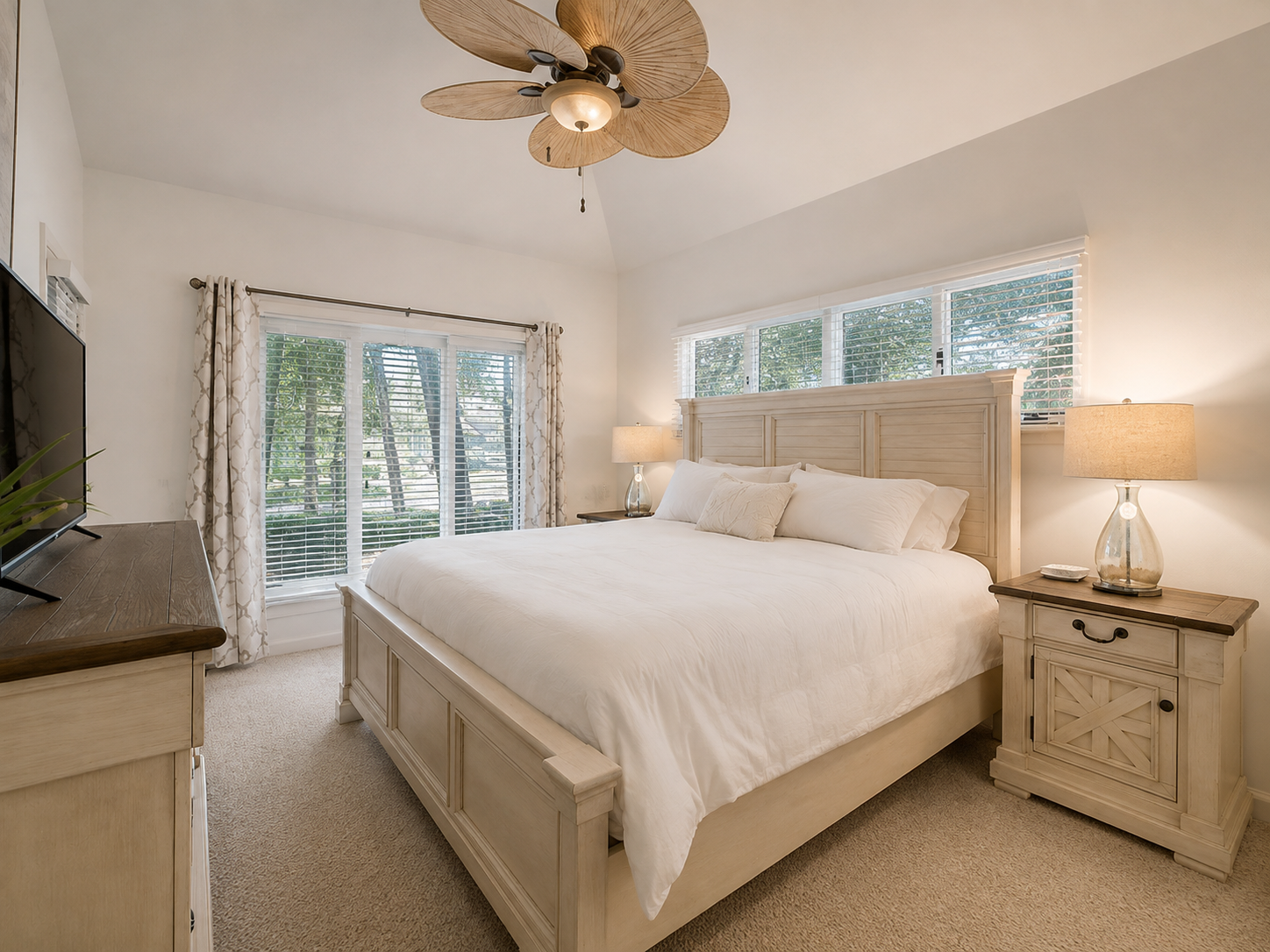 Bright bedroom with a large white bed, wooden nightstands with lamps, a wooden dresser with a TV, and large windows with blinds and patterned curtains, tropical ceiling fan.