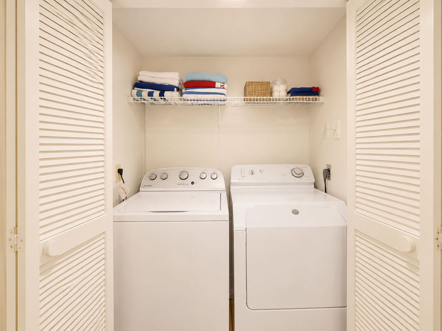 A laundry closet with a washing machine and dryer, white shelves holding folded towels and a basket, with louvered closet doors.