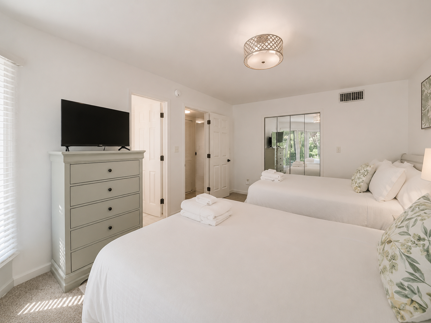 A bright, minimalist bedroom with two white beds, a gray dresser, a wall-mounted flat-screen TV, a mirrored closet door, and a window with blinds. The room has white walls and ceiling, with a modern light fixture.