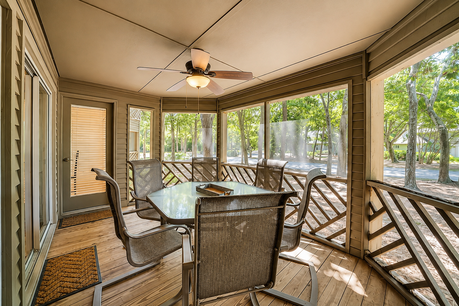 A screened porch with a wooden floor, ceiling fan, glass-top table, and six chairs, overlooking a yard with trees and a street in the background.