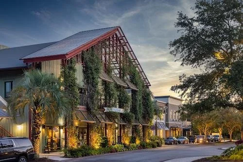 A two-story building with a red metal roof and wooden exterior, surrounded by palm trees and street parking at sunset.