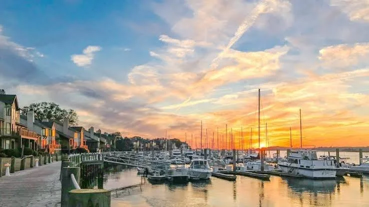 Sunset over a marina with boats docked and houses along the waterfront.