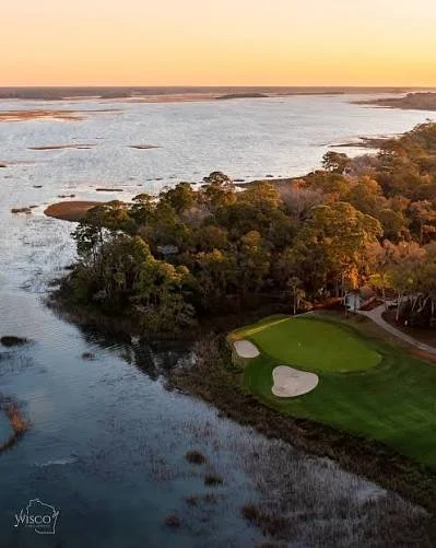 An aerial view of a river and a golf course at sunset, with trees along the shoreline and water bodies in the background.