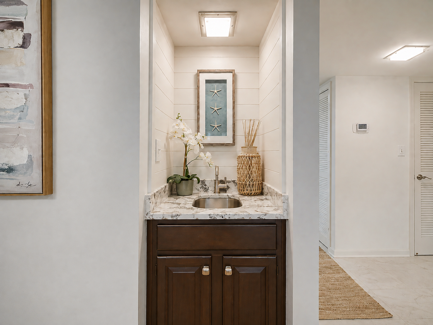 A small decorative wet bar with a marble countertop, a miniature sink, a potted orchid plant, two woven baskets, and a framed artwork with starfish, set against white shiplap paneling.
