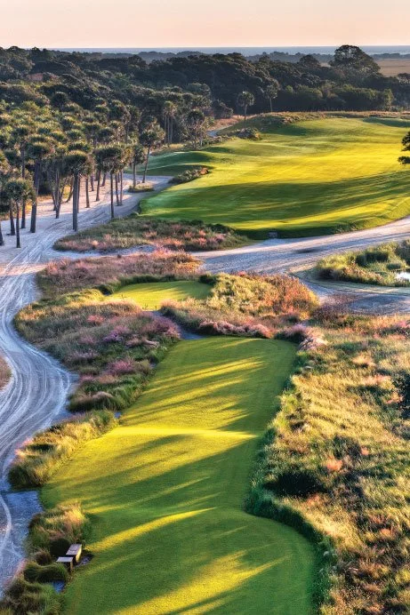 Aerial view of a golf course with green fairways, sand traps, and surrounding trees during sunset.
