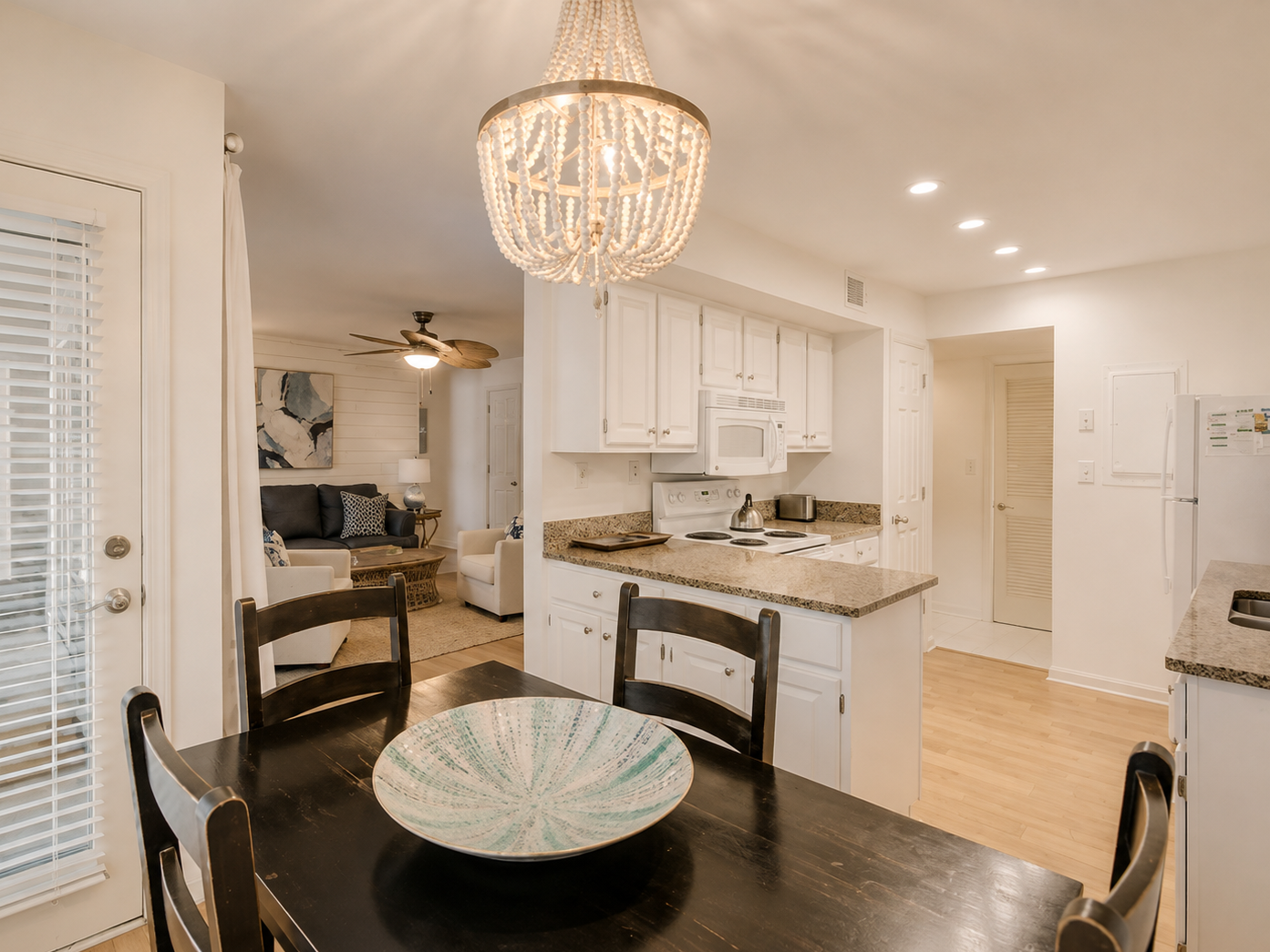 Dining table with a decorative bowl, kitchen with white cabinets, granite countertops, white appliances, and a chandelier, with a living room in the background.