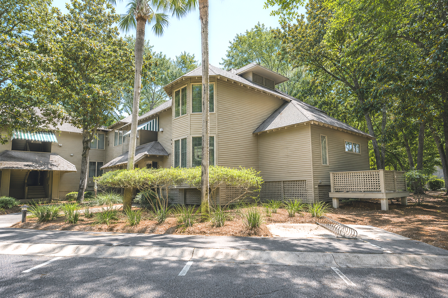 A beige multi-story residential building with a deck, surrounded by trees and landscaping, adjacent to parking spaces.