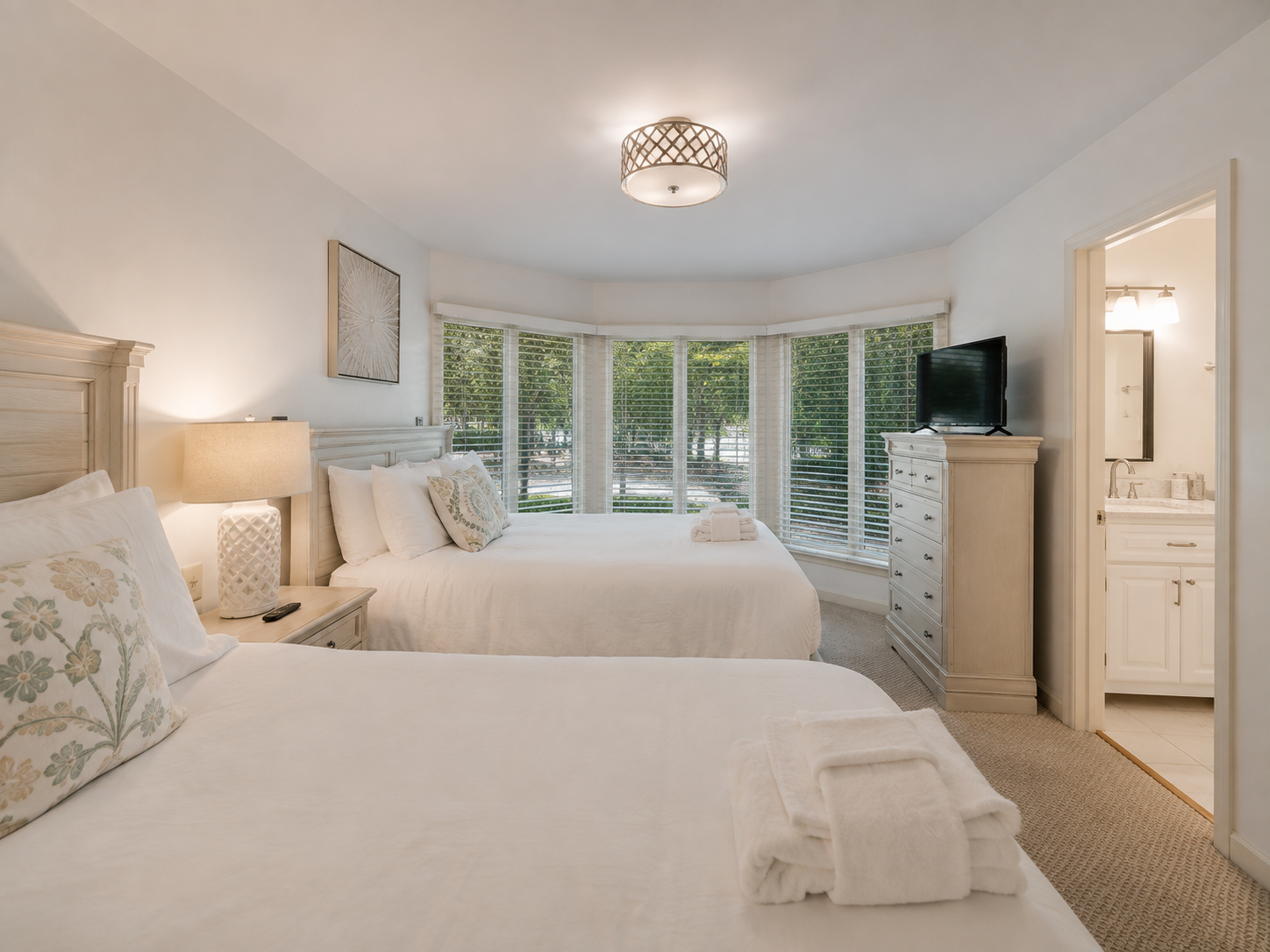 A bright hotel room with two white beds, a dresser with a TV on top, and a bay window overlooking green trees. A doorway to a bathroom is visible.