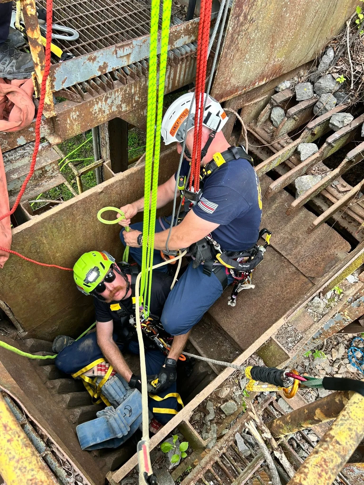 Rescue workers are working inside a rusted, exposed steel pipe or culvert, possibly performing a rescue or inspection. The scene includes safety ropes, helmets, and harnesses, indicating an industrial or construction environment.