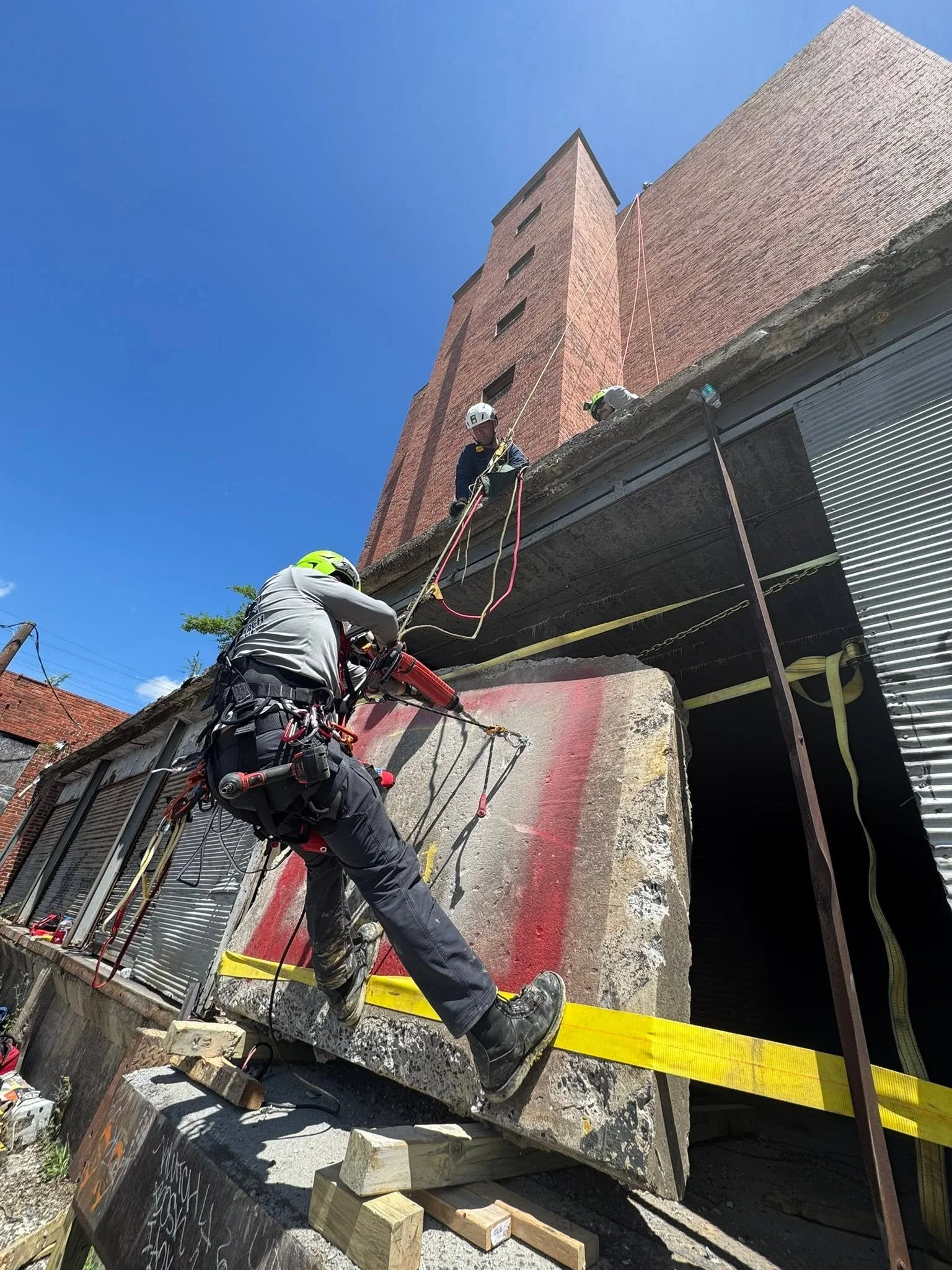 Workers in helmets and harnesses are performing demolition or construction work on a building's facade, with reinforcement equipment and debris visible around.